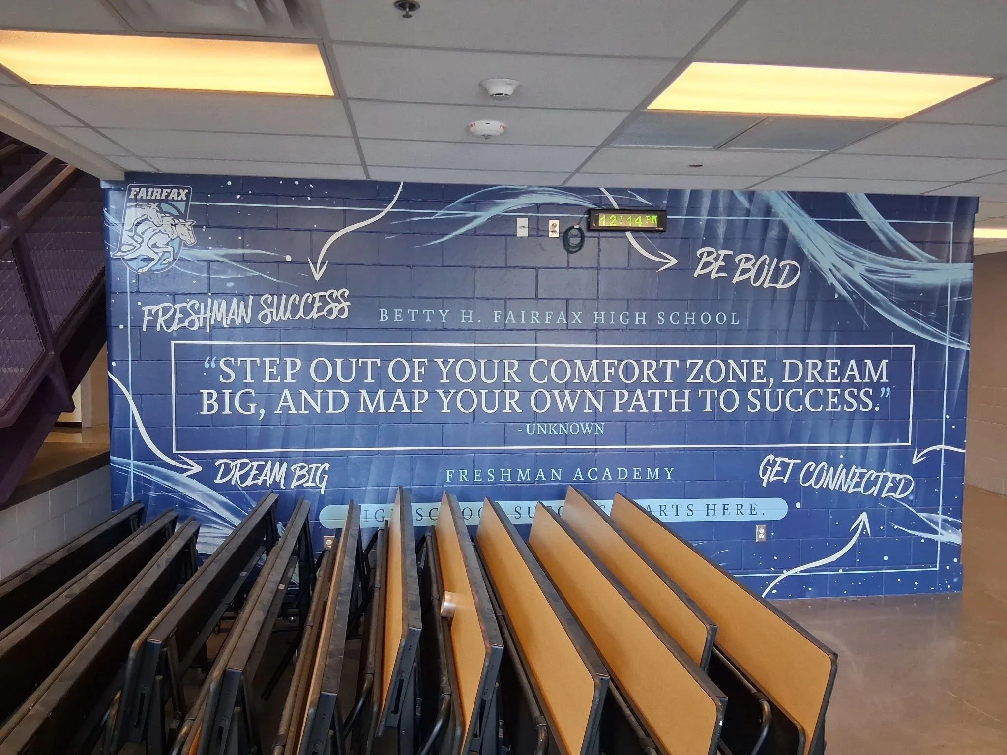 A staircase leading up to a large blue banner on the wall. The banner features the Fairfax logo with a horse and rider, inspirational quotes, and educational mottos such as "Freshman Success" and "Be Bold." The time on a digital clock reads 12:14 PM.
