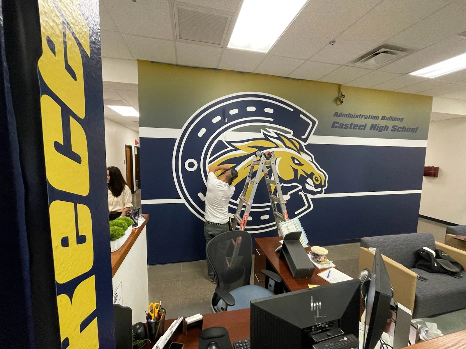 A man is standing on a ladder working on a large mural of a school mascot, a horse, inside an administration building of Casteel High School. There is a woman sitting at a desk nearby, and other office supplies and furniture are visible in the room.