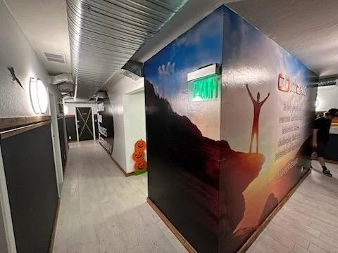 Hallway with a wall mural of a person with arms raised on a mountain at sunset, and a small scarecrow figure at the corner.