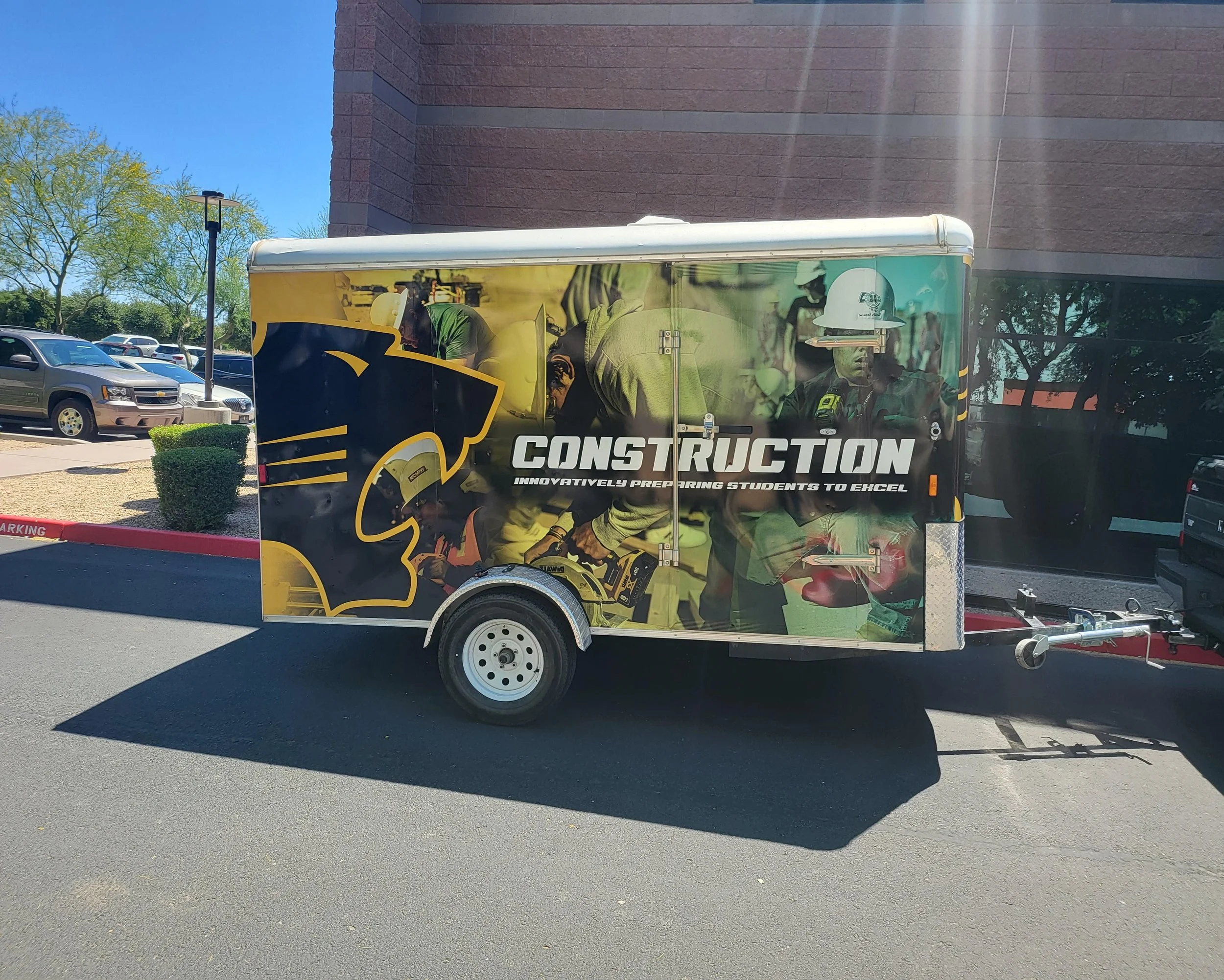 A trailer with construction-themed graphics parked in a lot, with other cars and trees visible in the background.
