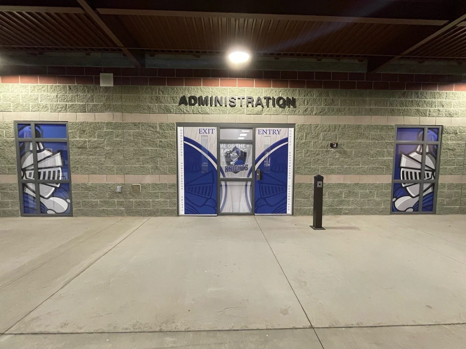 The entrance to an administration building decorated with blue and white sports-themed graphics featuring a knight mascot with the text "Knights". The building has two windows with similar sports graphics and a door with the same mascot. Above the do