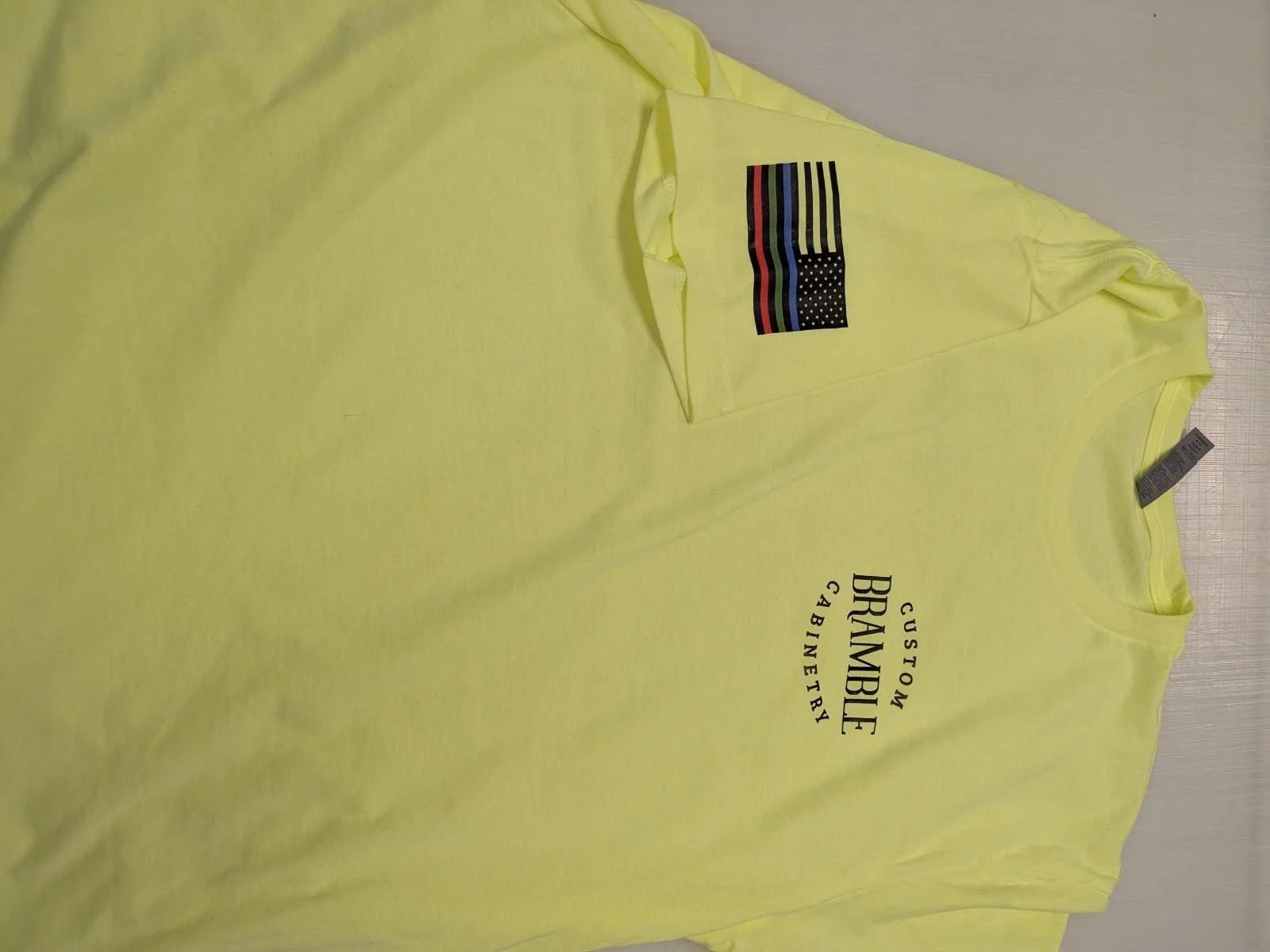 Yellow T-shirt with a graphic design and text that reads 'Custom Bramble Cabinetry' and an American flag patch on the chest.