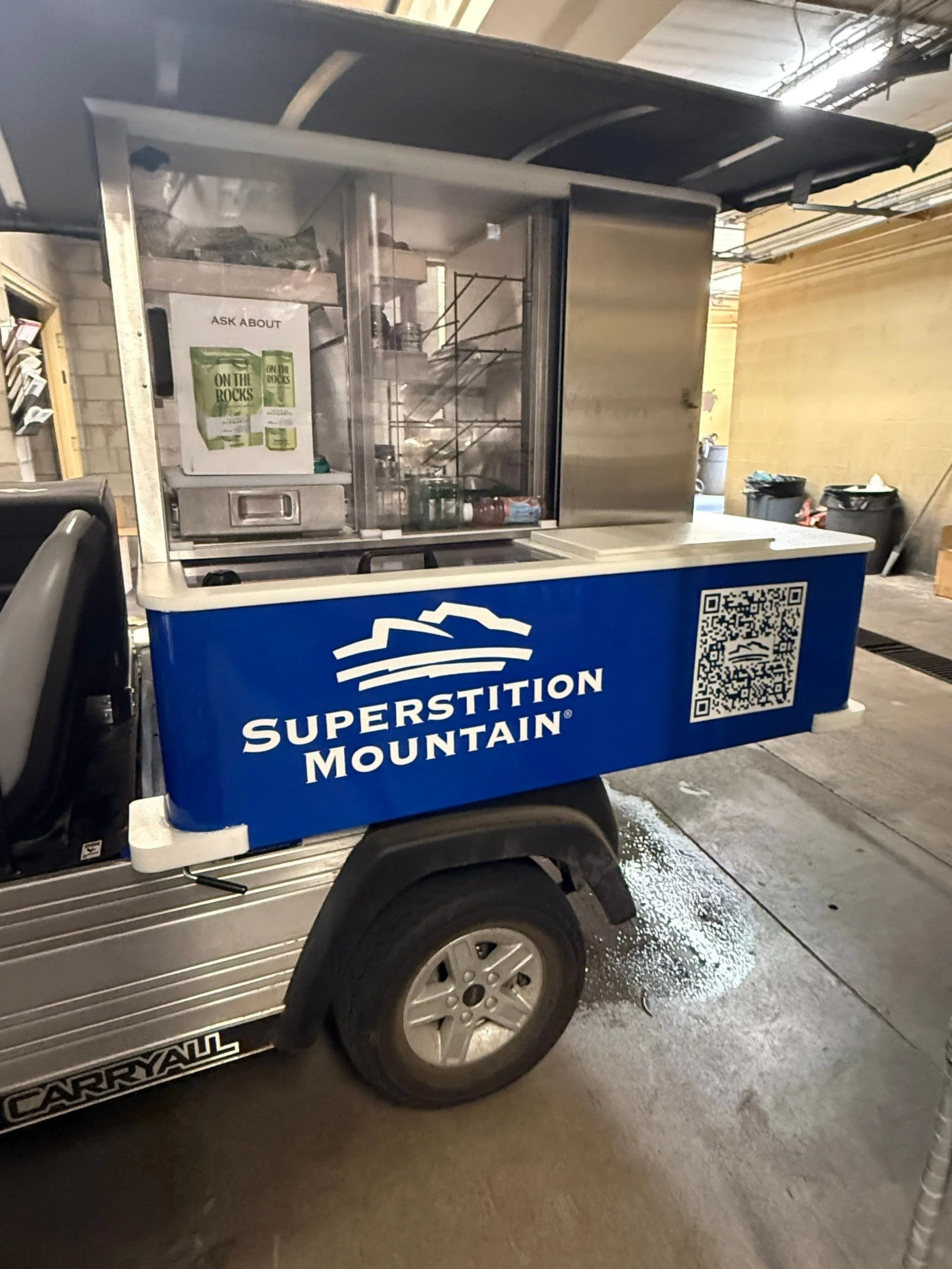Close-up of a Superstition Mountain branded golf cart or utility vehicle with a promotional sign for a product called 'On The Rocks' inside the clear display case. The vehicle is parked inside a garage or warehouse with recycling bins and various obj