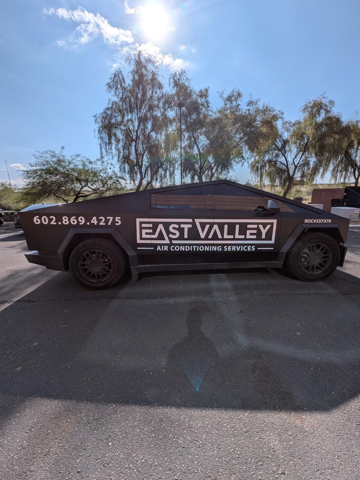 Black car with the logo and phone number for East Valley Air Conditioning Services printed on the side, parked on an asphalt lot with trees and a blue sky with the sun overhead.