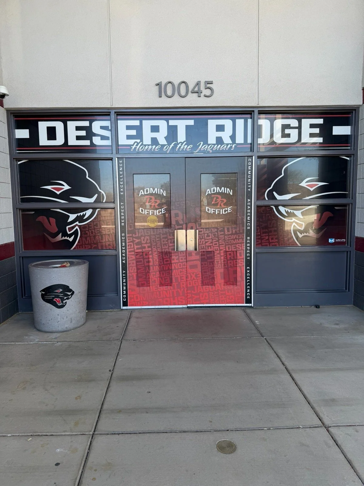 School entrance with glass doors decorated with jaguar logos, the name 'Desert Ridge' and the phrase 'Home of the Jaguars,' along with words like respect, excellence, community, academics, and pride. A trash bin with a jaguar logo is on the left, and