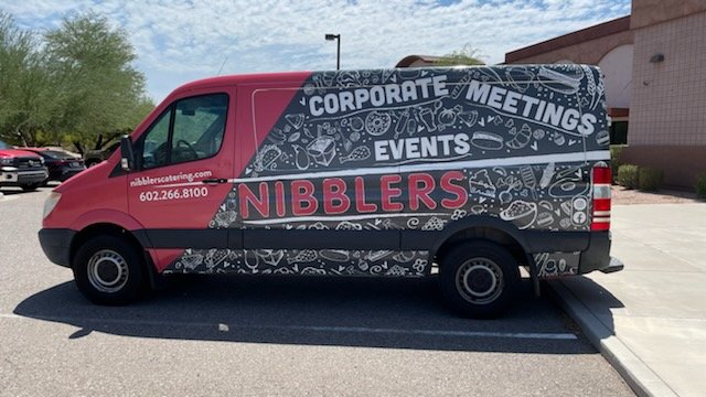 A pink and black van with professional graphics advertising Nibbler's catering services, including corporate meetings and events, parked in a parking lot.