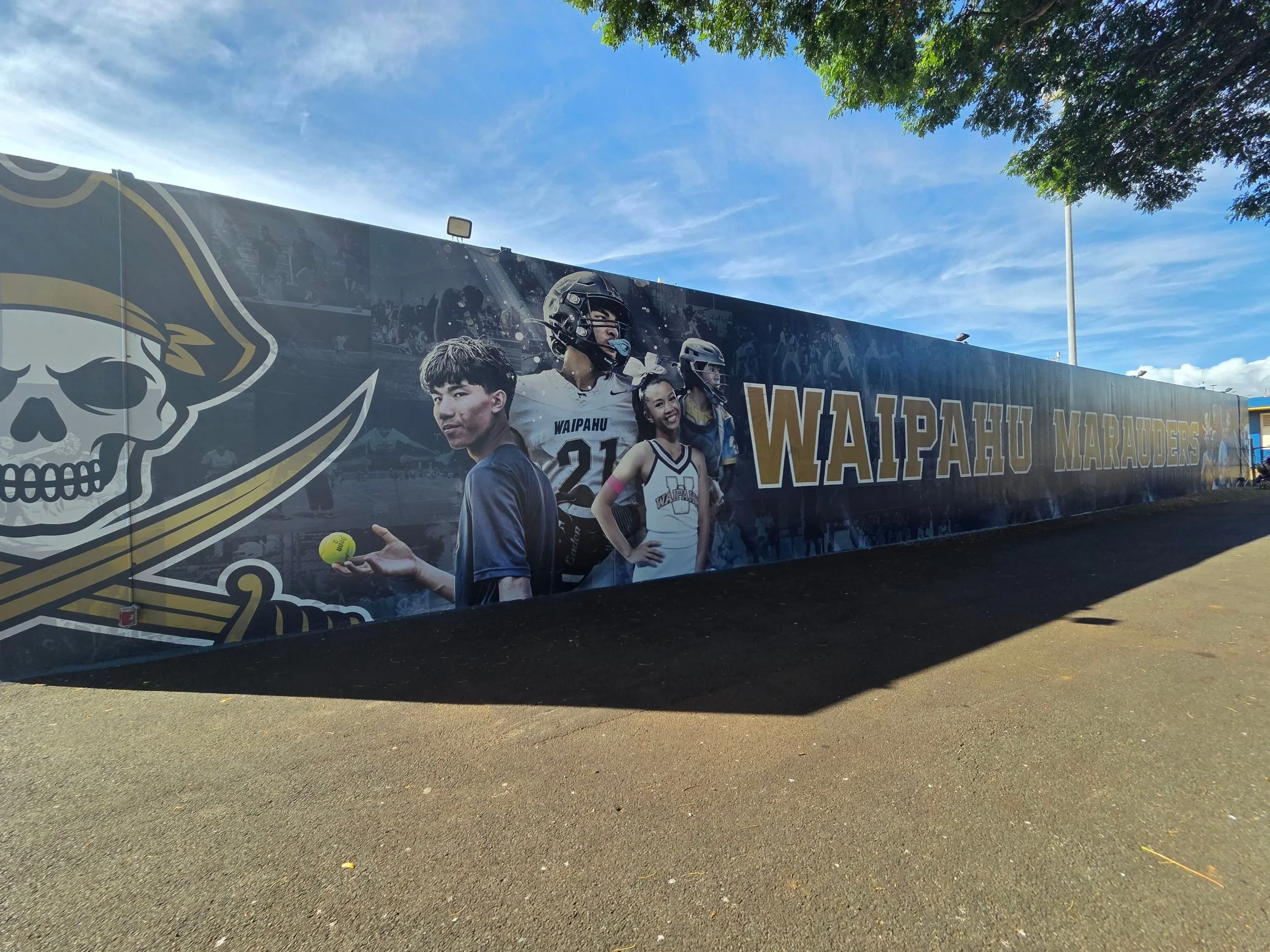 A large mural mural featuring sports players and a prominent skull logo with a crown. The mural is displayed outdoors on a low wall with a clear blue sky and a few trees in the background. The words "Waipahu Marauders" appear on the mural.