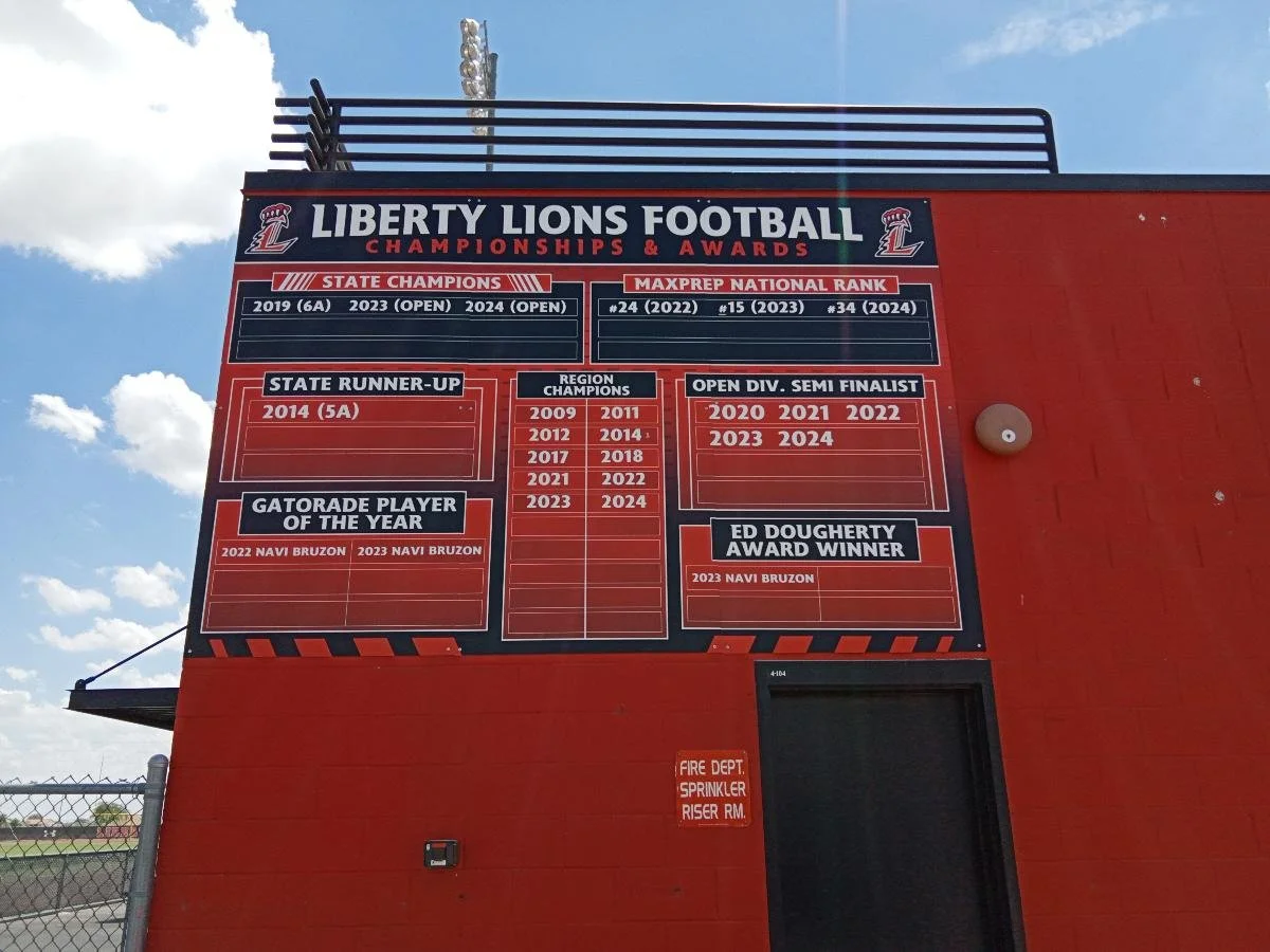 Red scoreboard on a red wall at Liberty Lions football field displaying team achievements and awards, including state championships, regional champions, and individual awards.