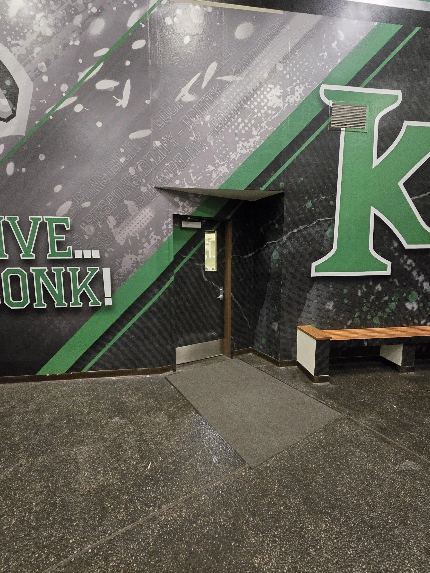 A wall with a sports-themed mural featuring a large green and white letter 'K'. The mural includes words like 'ESPBILITY', 'EDUCITY', and 'KINDNESS' along with various paint splatters and design elements. There is a small ventilation grate and a wood