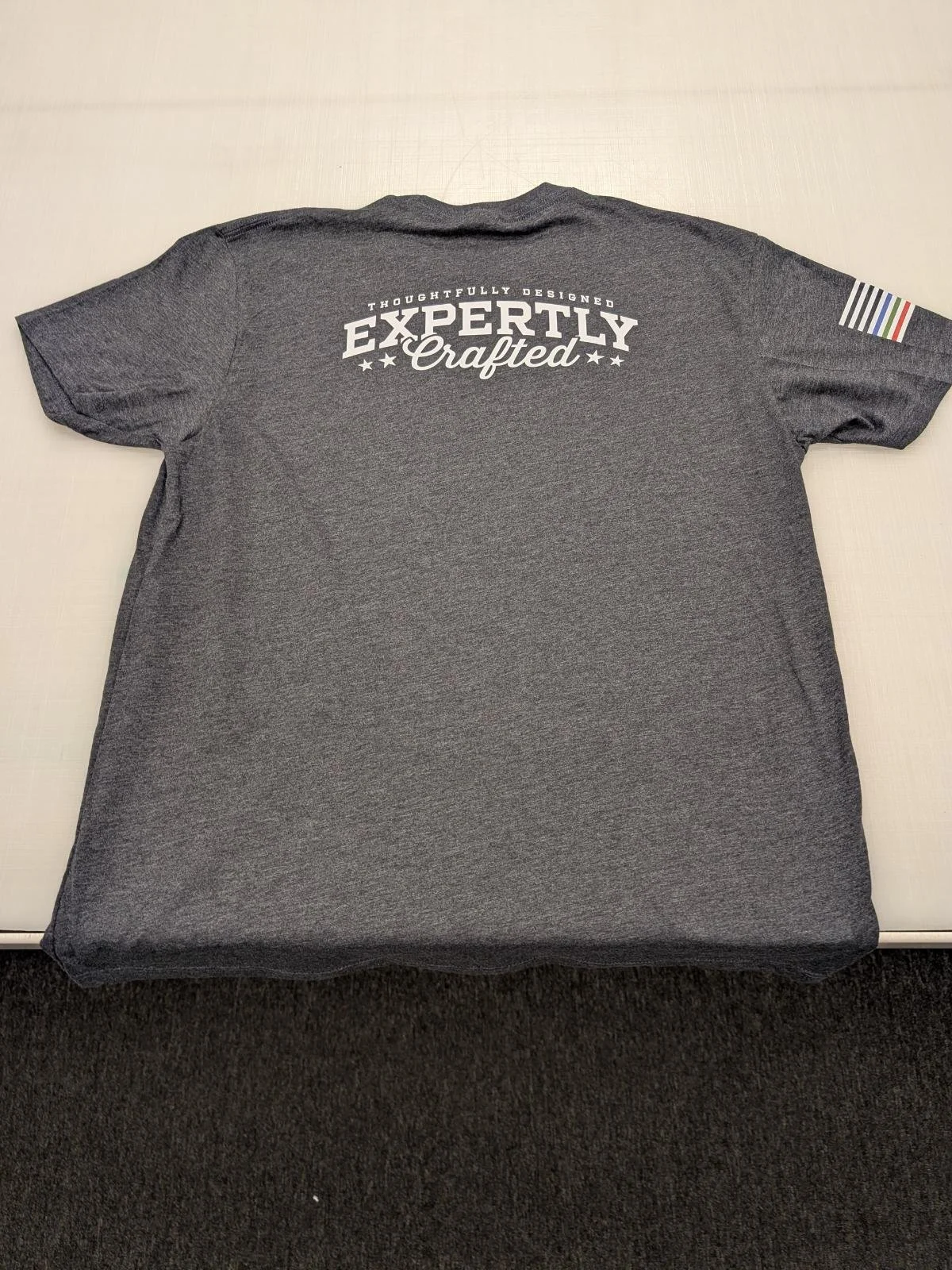 Gray T-shirt with white text on the back that says "Thoughtfully Designed Expertly Crafted" and an image of stars, with a striped pattern on the right sleeve.