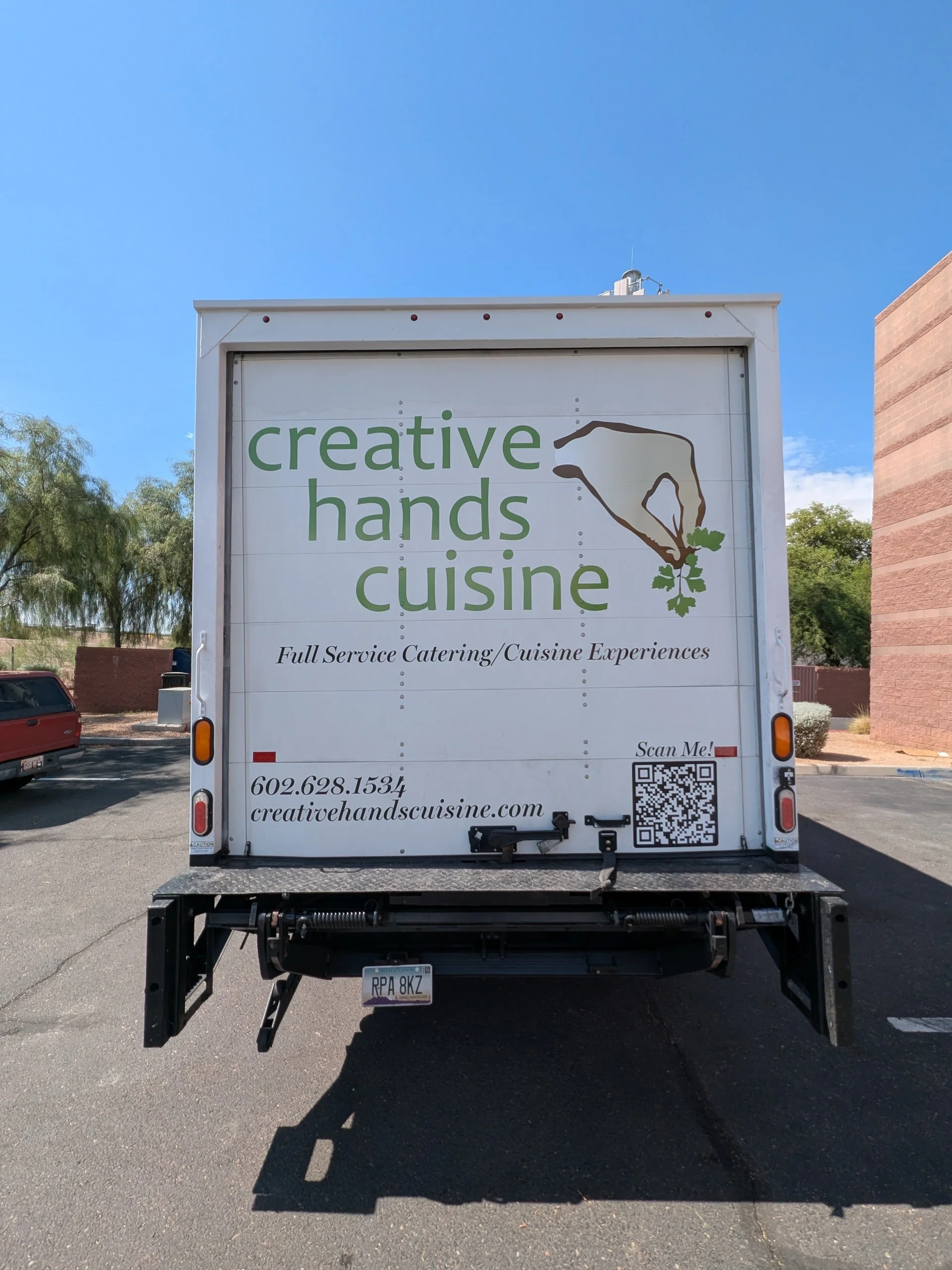 The back of a white catering truck with the logo 'creative hands cuisine' in green and brown, featuring a hand drawing herbs. The truck provides full-service catering and cuisine experiences, and includes a QR code and contact information.