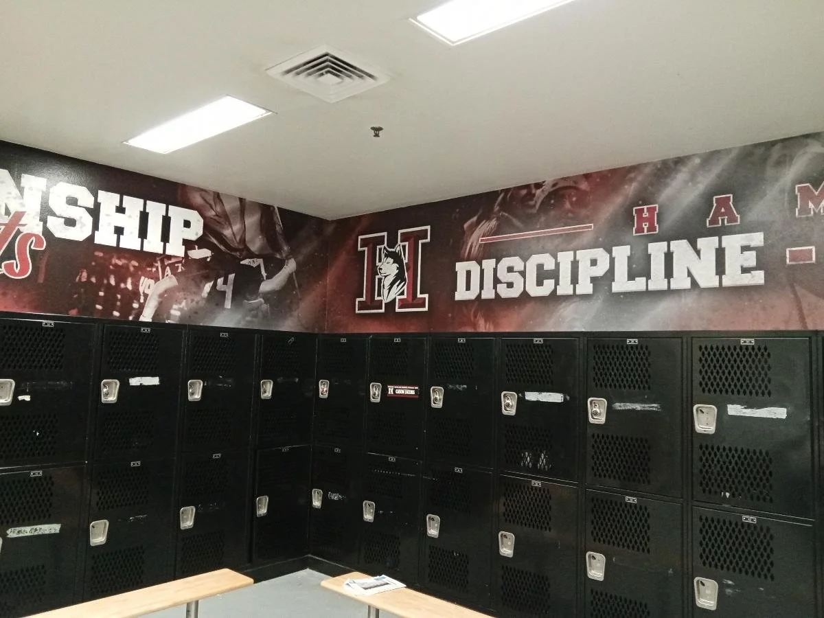 Locker room with black lockers and a mural on the wall featuring athletic imagery and the words "Discipline" and other sports-related text.