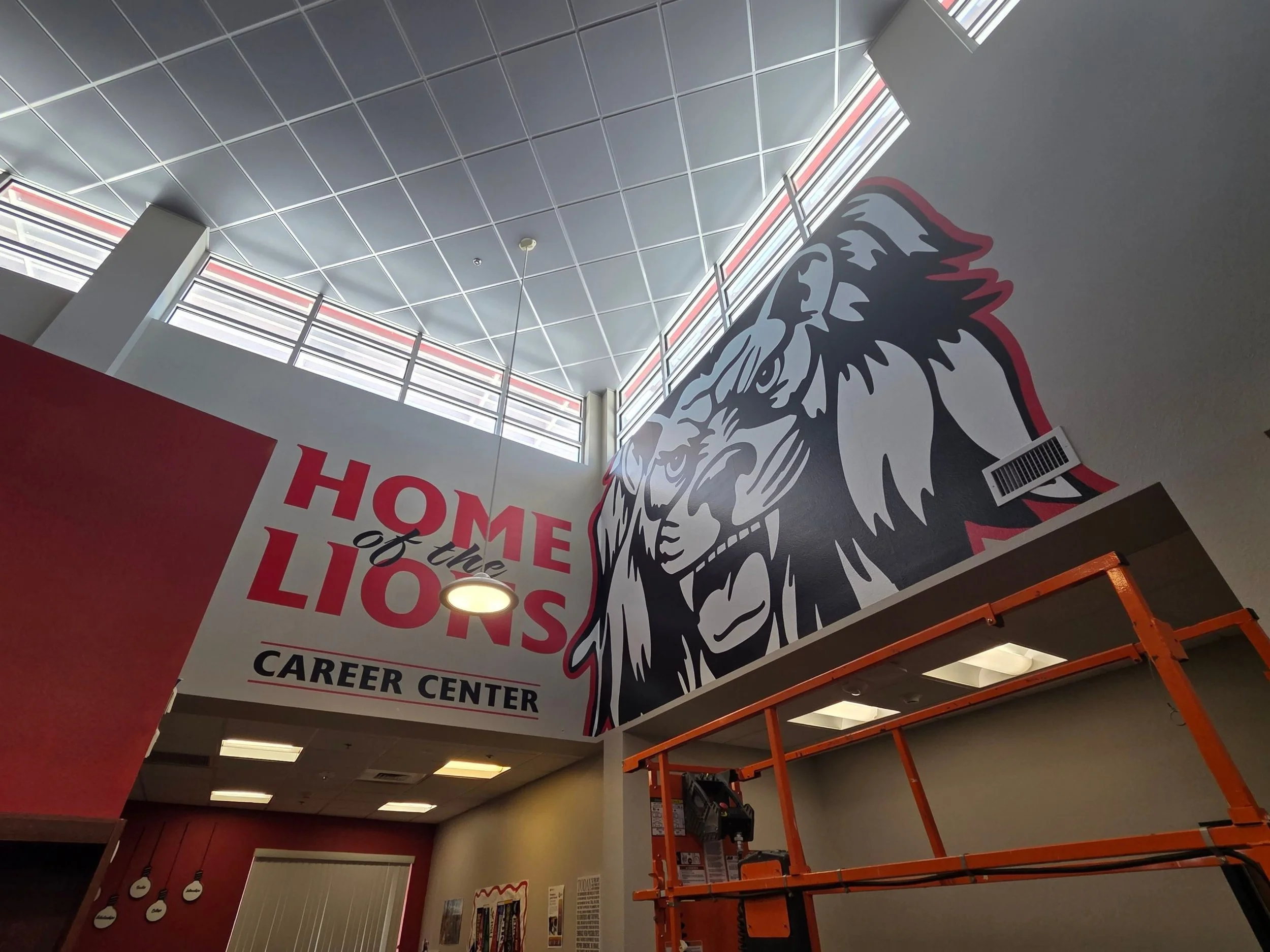 Interior of a building with large windows on the ceiling, revealing a red and white striped pattern outside. There is a portion of a large lion graphic on the wall with the words 'Home of the Lions Career Center' partially visible. An orange scaffold