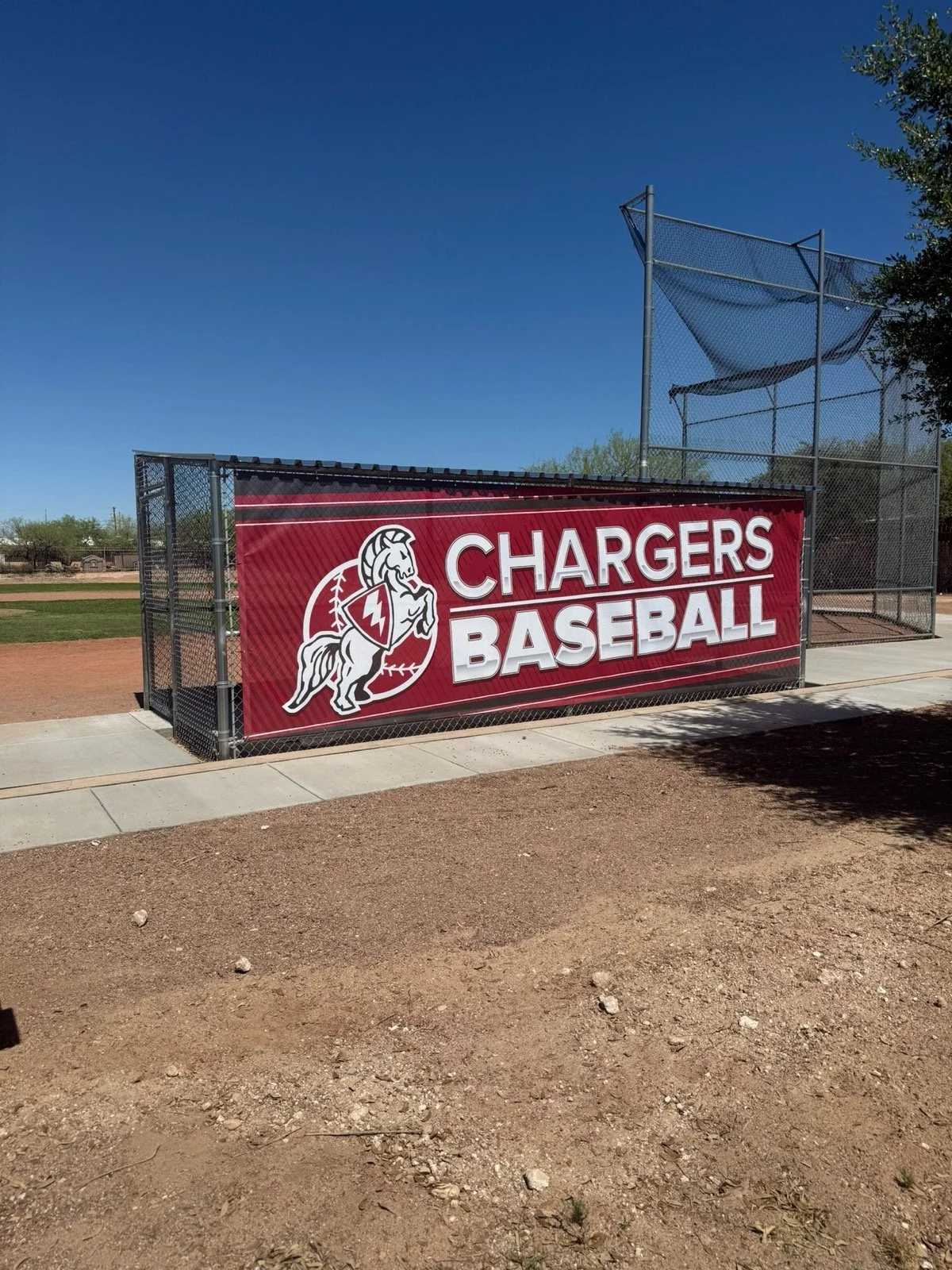 Baseball dugout with a red banner that reads 'Chargers Baseball' and features a logo of a charging horse with a shield. The dugout has a chain-link fence, and a baseball field is in the background under a clear blue sky.