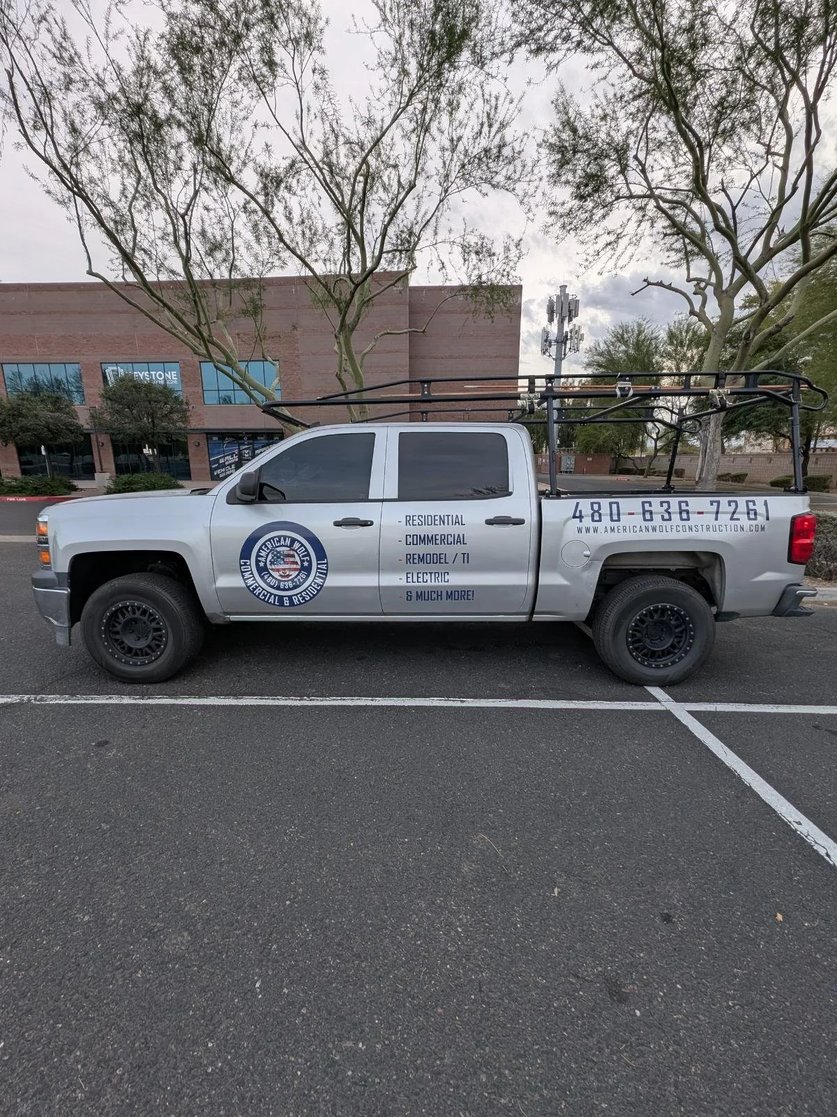 Silver pickup truck parked in a parking lot with service advertisements on the side, including company name, phone number, and services offered, such as residential, commercial, remodel, electrical, and more, with a ladder rack on top and a building 