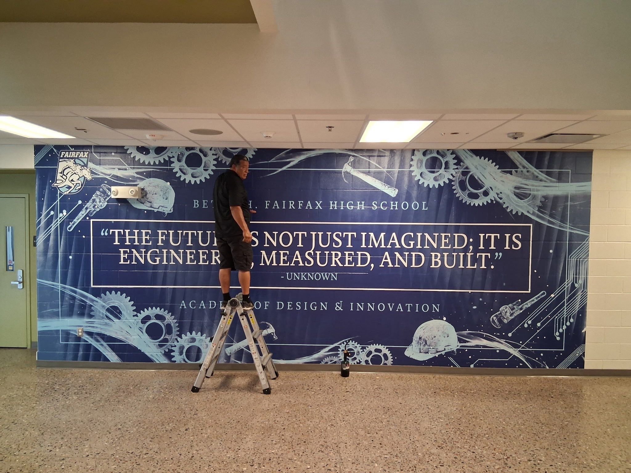 A man standing on a step ladder working on a large blue poster with gears, mechanical sketches, and a quote, in a hallway.