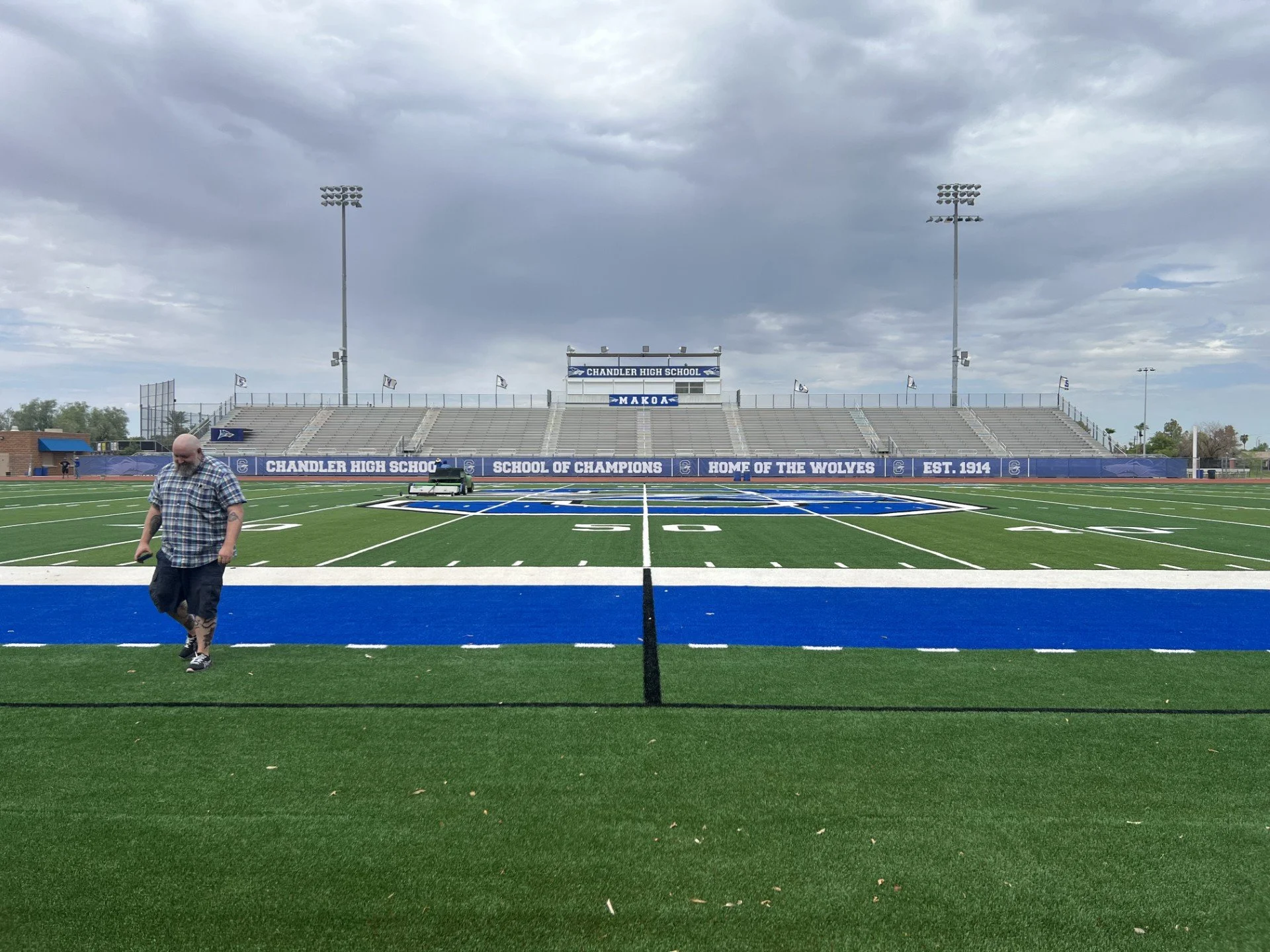 Football field with bleachers, flags, and sign reading 'Chandler High School', under cloudy sky, with a man walking on field.