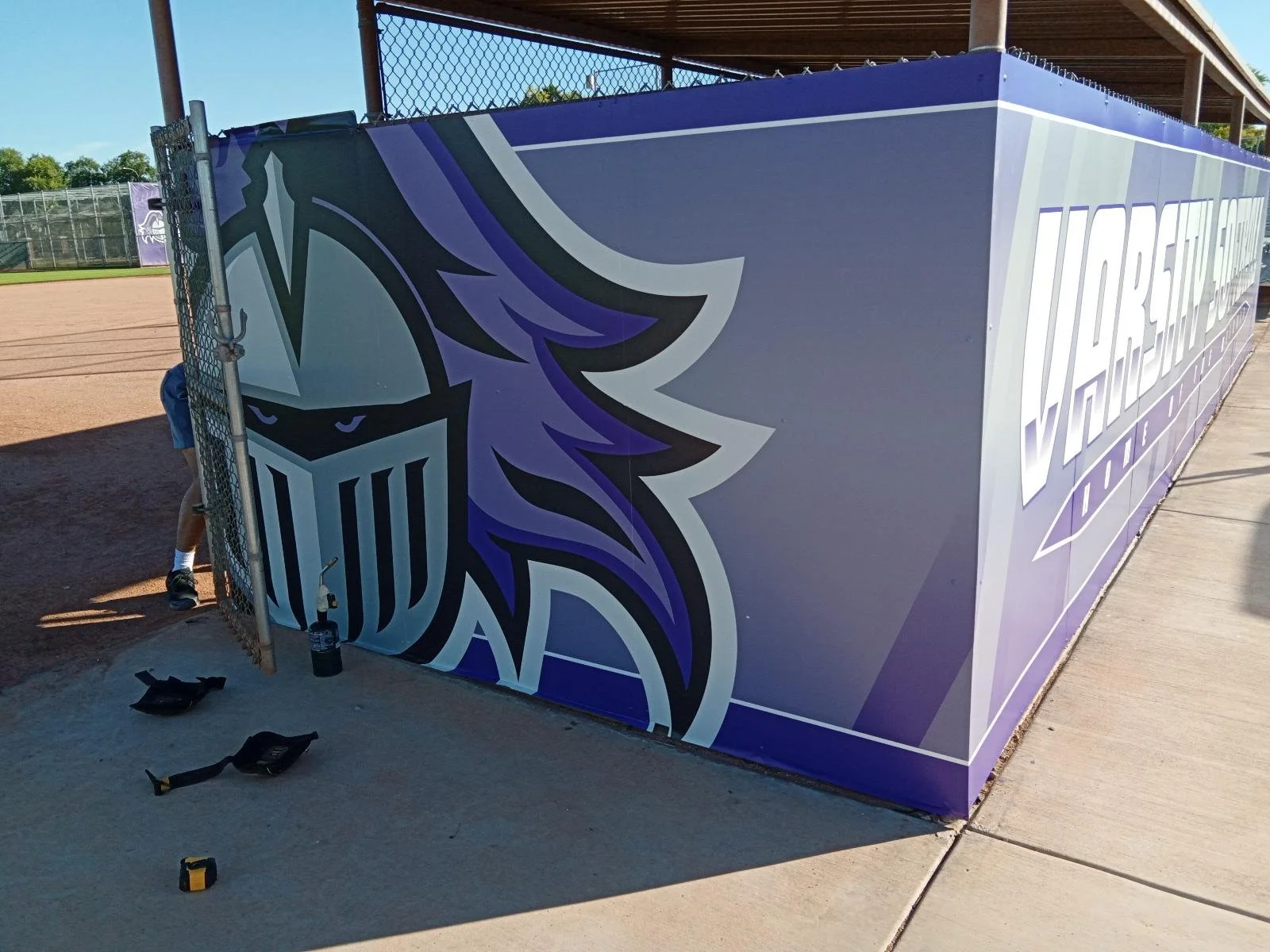 A purple Denver Broncos barrier with the Broncos logo, featuring a stylized horse head with a flowing mane and a shield, on a baseball field dugout.