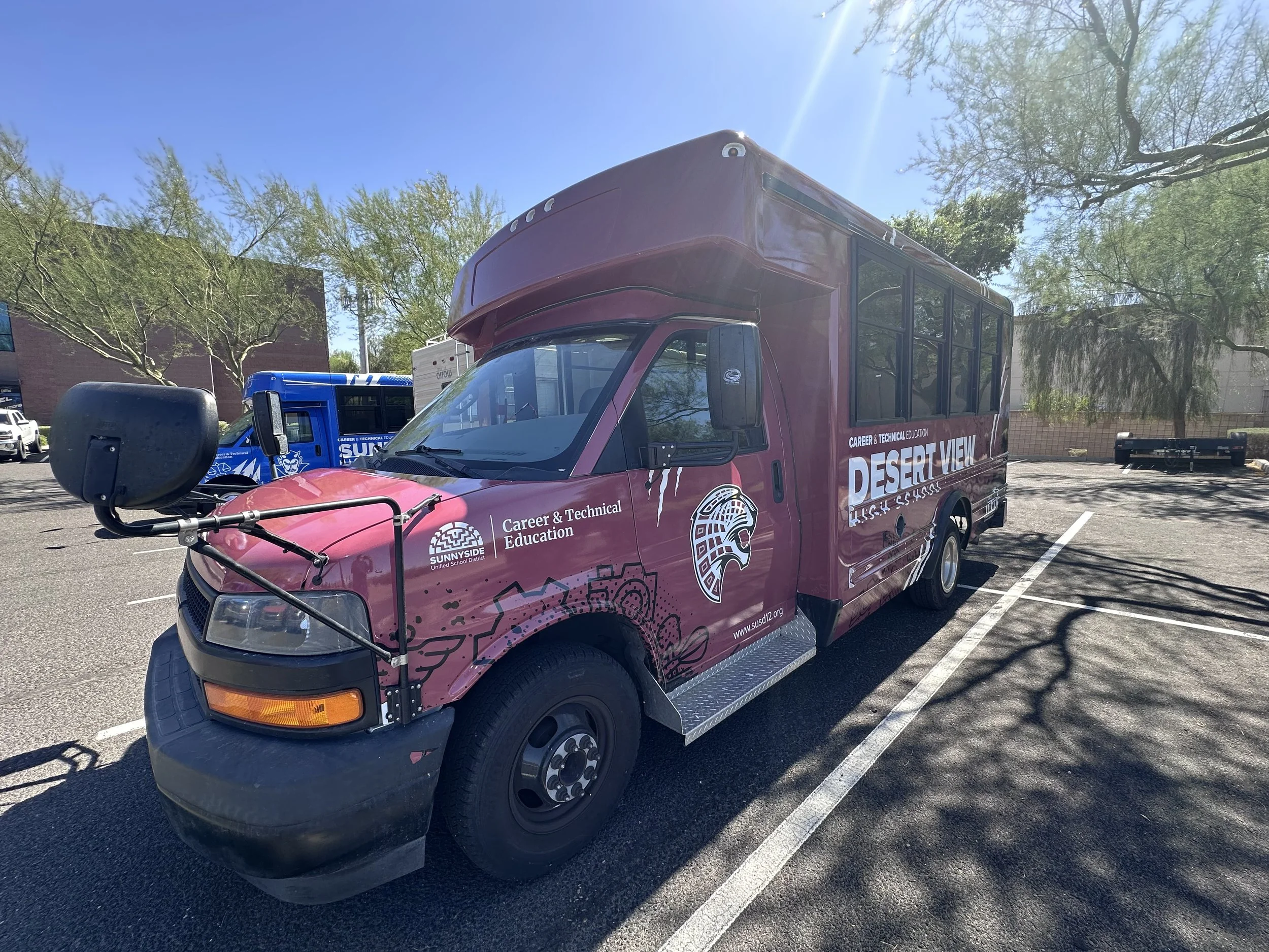 A maroon van with the words "DESERT VIEW" and "CAREER & TECHNICAL EDUCATION" on the side, parked in a lot with trees and blue sky.
