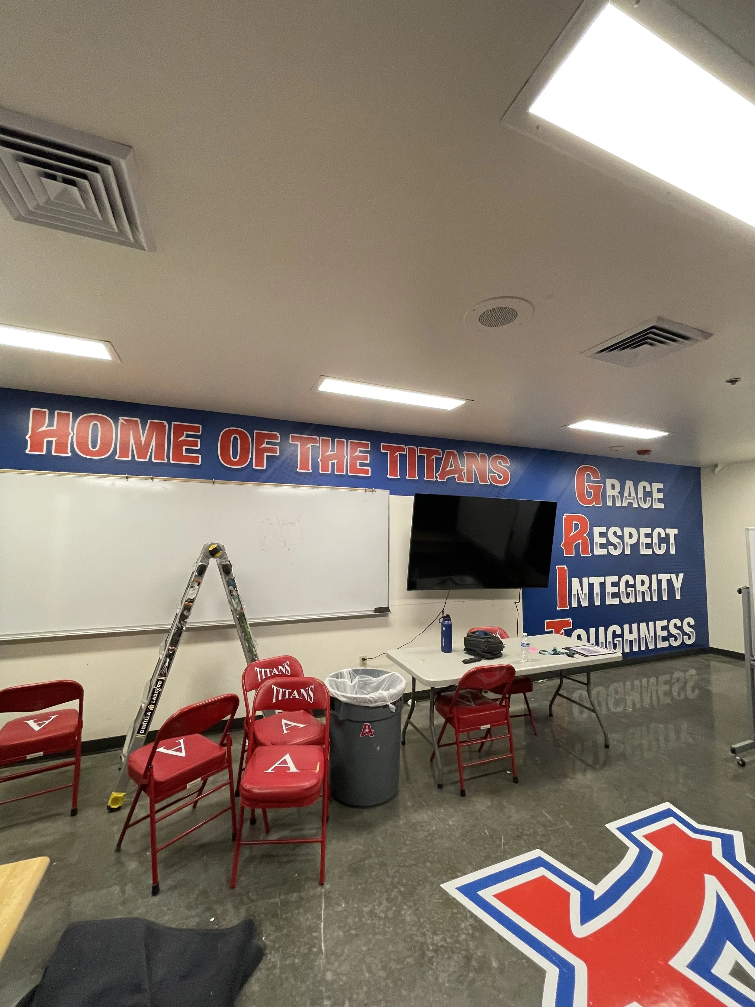 A classroom or meeting room decorated with a large sign that reads 'Home of the Titans' and features a list of school values or mottos such as 'Grace,' 'Respect,' 'Integrity,' and 'Toughness.' There are red chairs, a whiteboard, a ladder, a trash can