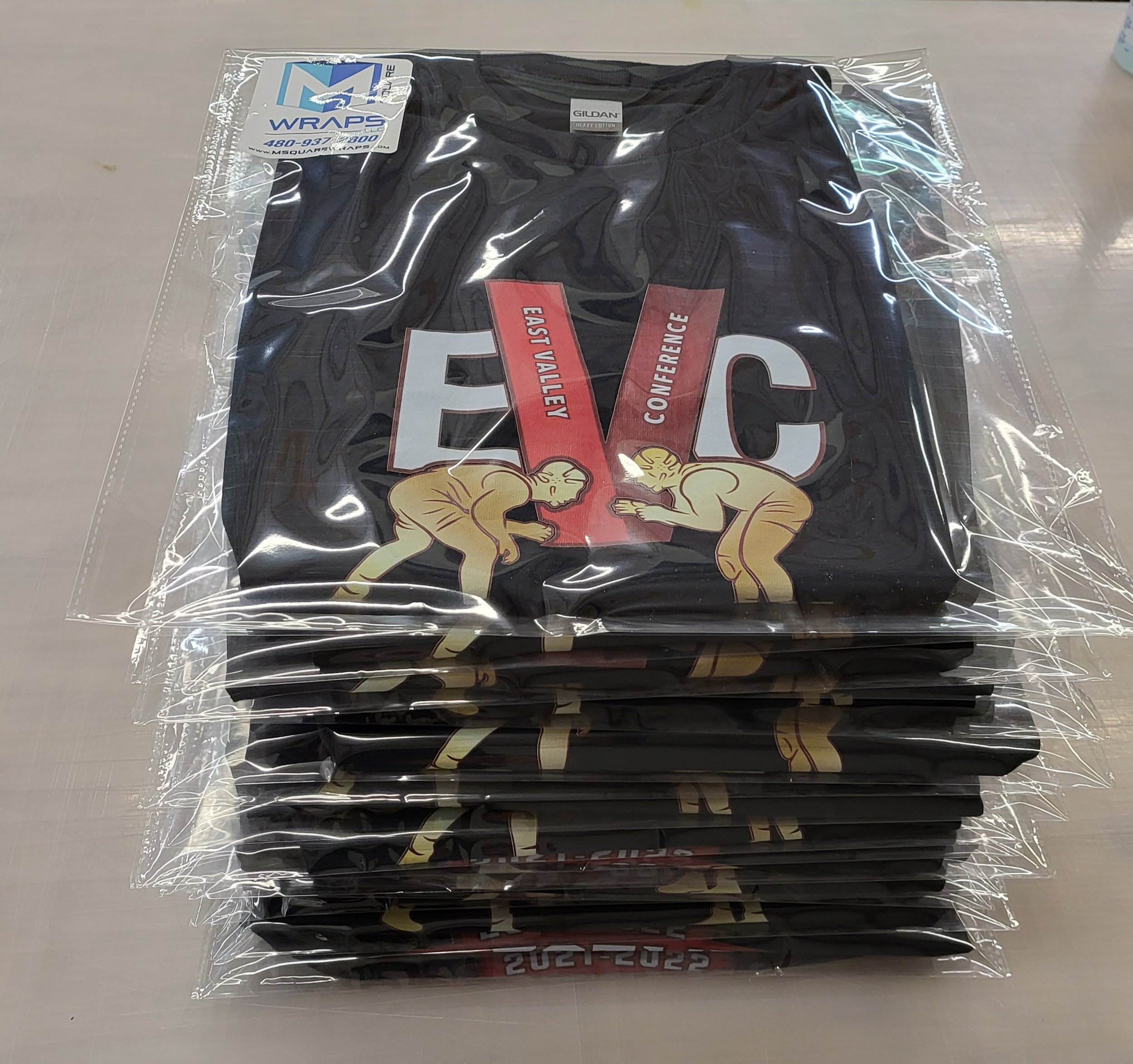 A black T-shirt inside clear plastic packaging with a label indicating it is heavy cotton, featuring a graphic of an arm wrestling match between two muscular men and text that includes the words 'EAST VA CONFERENCE'.