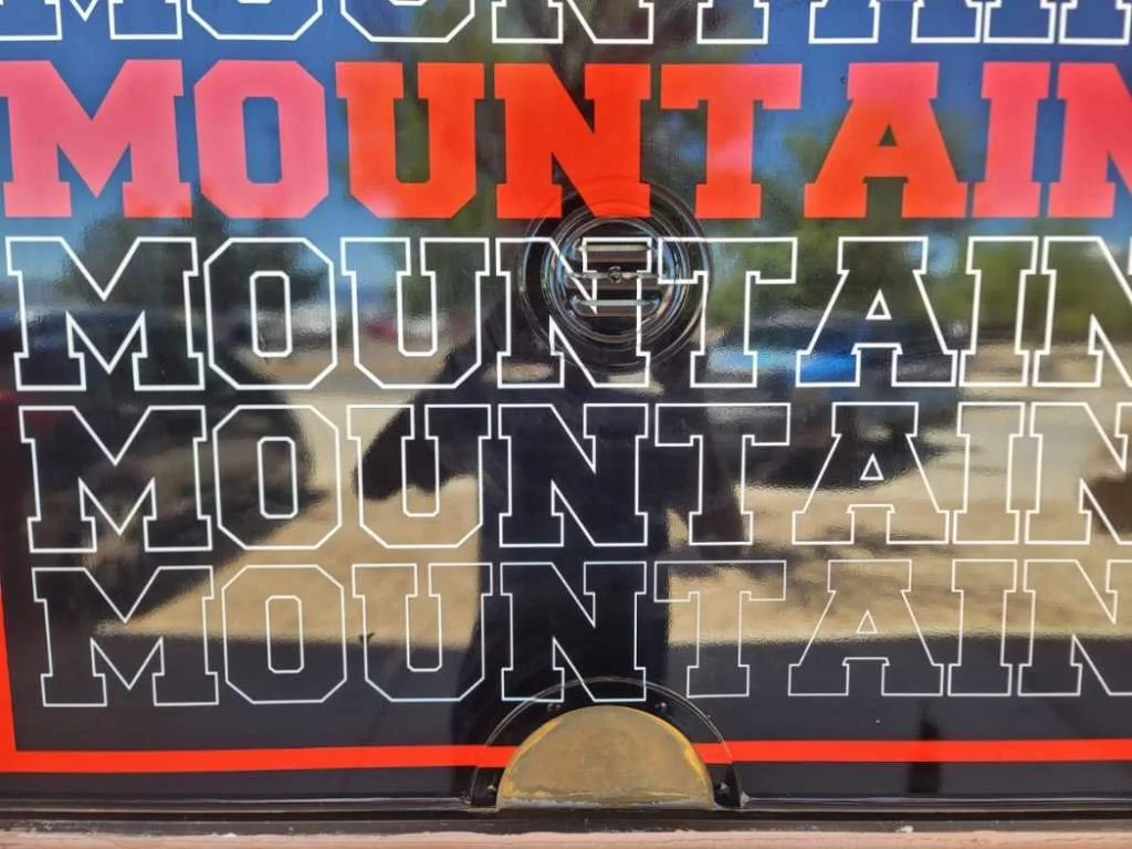 Reflection of sky and trees on a reflective surface with text 'MOUNTAIN' repeated multiple times and a camera lens in the center.
