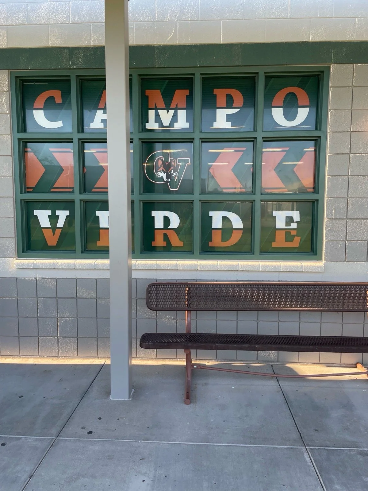 A window with the words "CAMPOS VERVE" in large colorful letters, with a logo of a wildcat's face in a circle in the middle. A black metal bench is in front of the window, and there is a vertical pole partially obstructing the view. The scene is outs