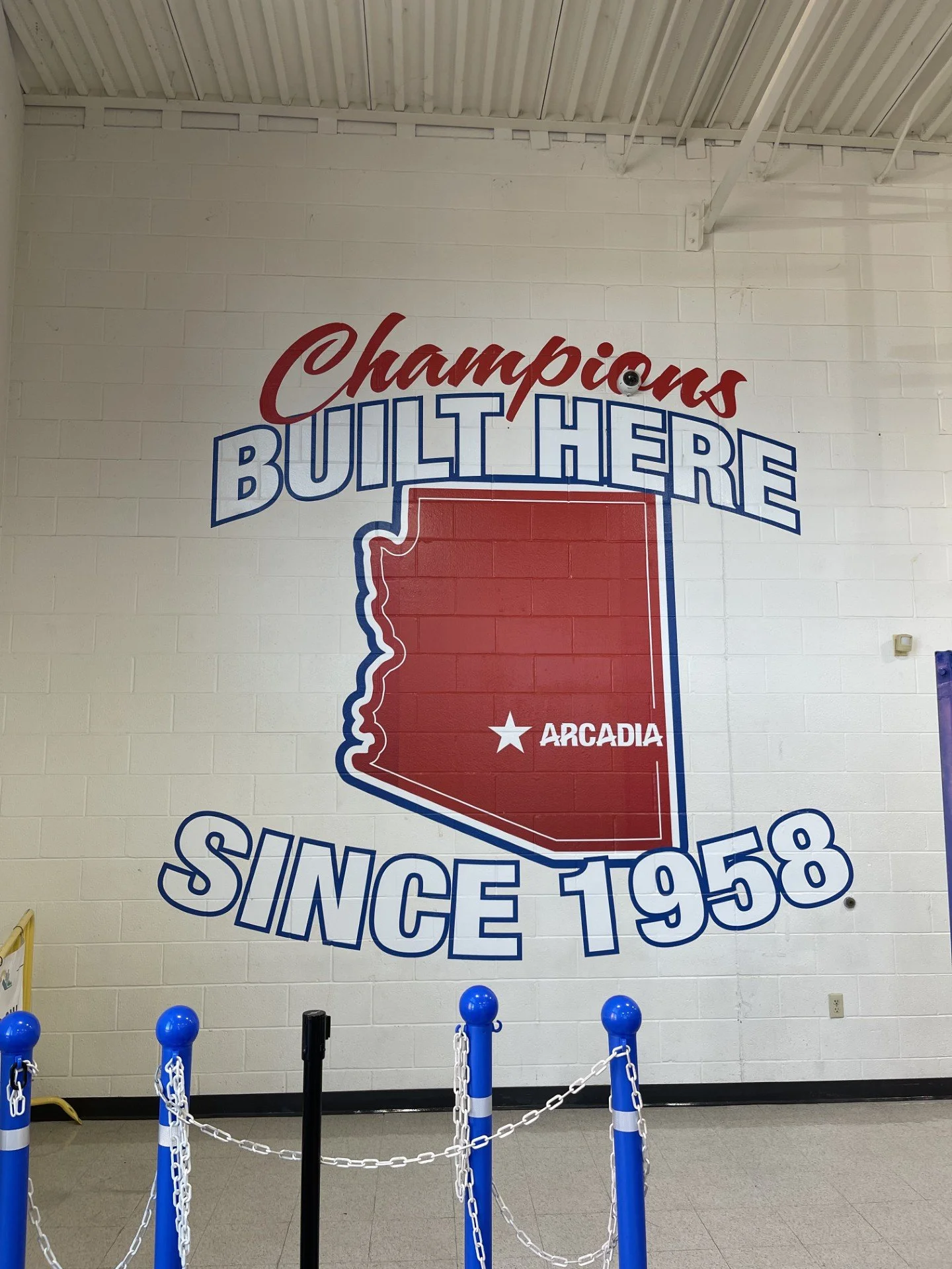 A mural on a white brick wall with the words "Champions Built Here" and "Since 1958". The mural includes a red outline of the state of Arkansas with the word "Arcadia" inside it. Blue and white chain barriers are in front of the mural, with blue post