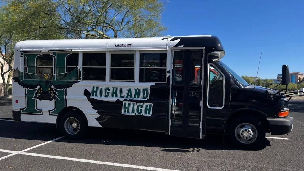 Black and white school bus with Highland High School logo featuring an eagle mascot, parked in a parking lot with trees and a building in the background.