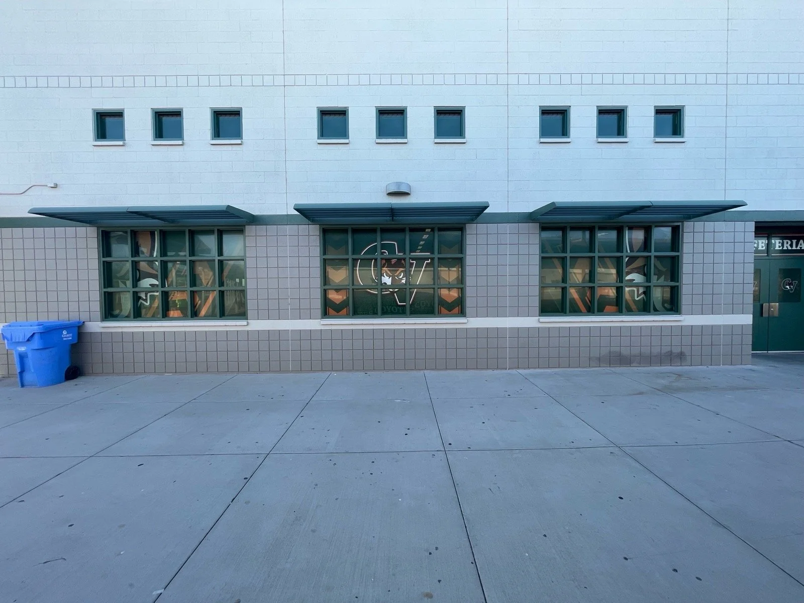 Exterior view of a building with three large windows with green frames, a smaller window with a logo, two awnings, a blue trash bin, and a gray sidewalk.