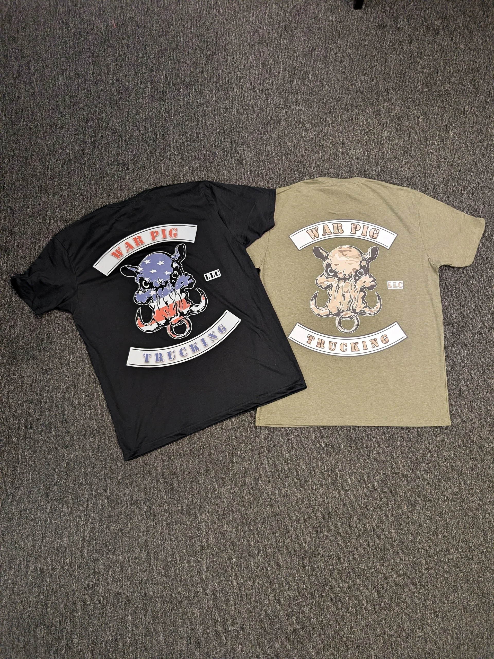 Two t-shirts, one black and one beige, with graphic designs of a pig's face and the words "WAR PIG TRUCKING" on the front, laid flat on a gray carpeted floor.
