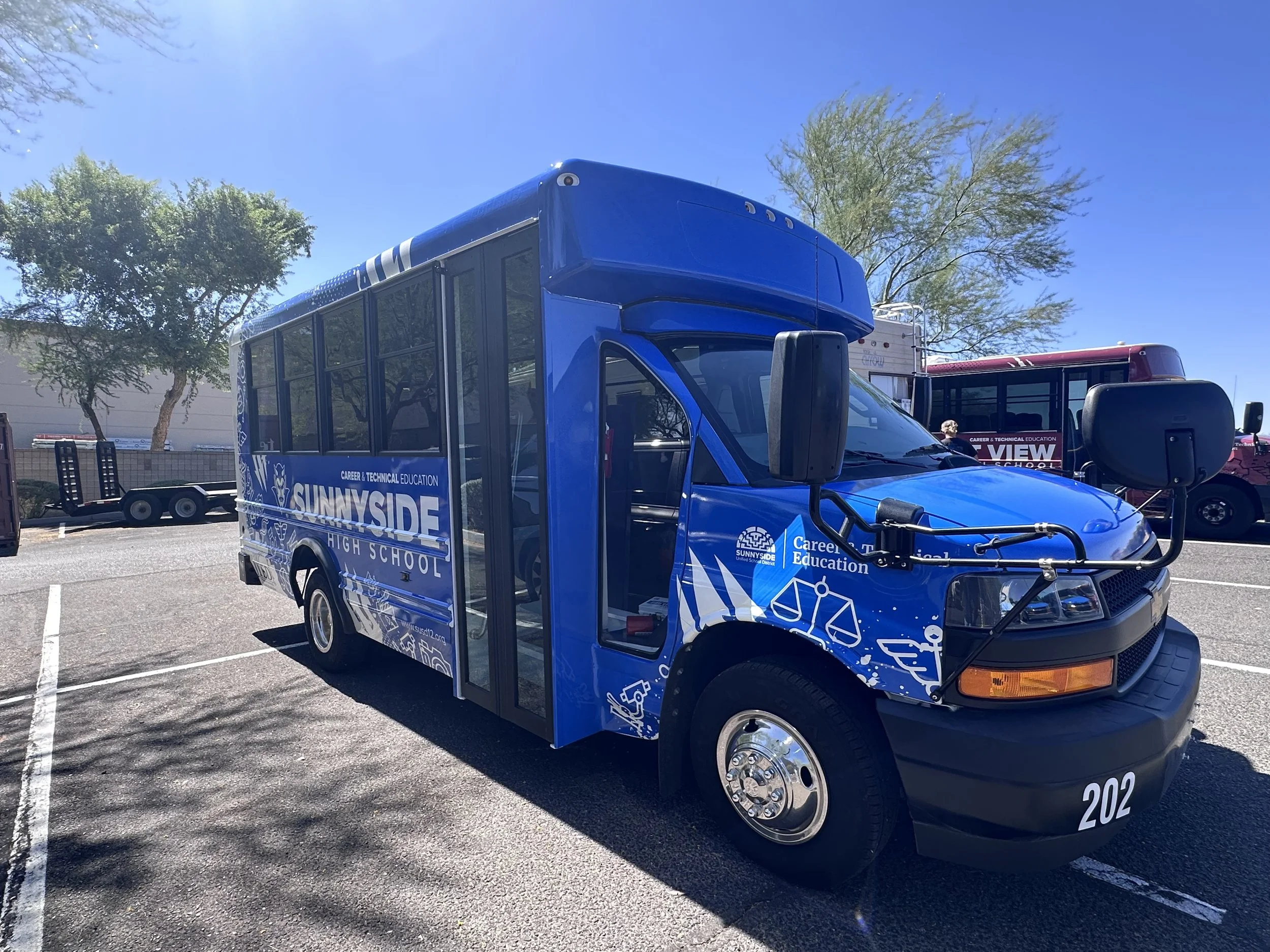 Blue mobile classroom vehicle with 'SUNNSIDE HIGH SCHOOL' and 'CAREER & TECHNICAL EDUCATION' written on the side, parked in a parking lot with trees and a clear blue sky in the background.