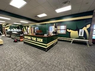 Interior of a museum exhibit area called 'Behind the Den' with a counter, seating, and a large wall decoration featuring a bull logo.