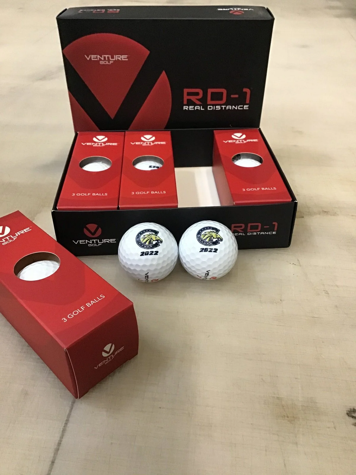 Box of Venture golf balls, with three individual golf balls outside the box, showing a logo with a lion and the year 2022.