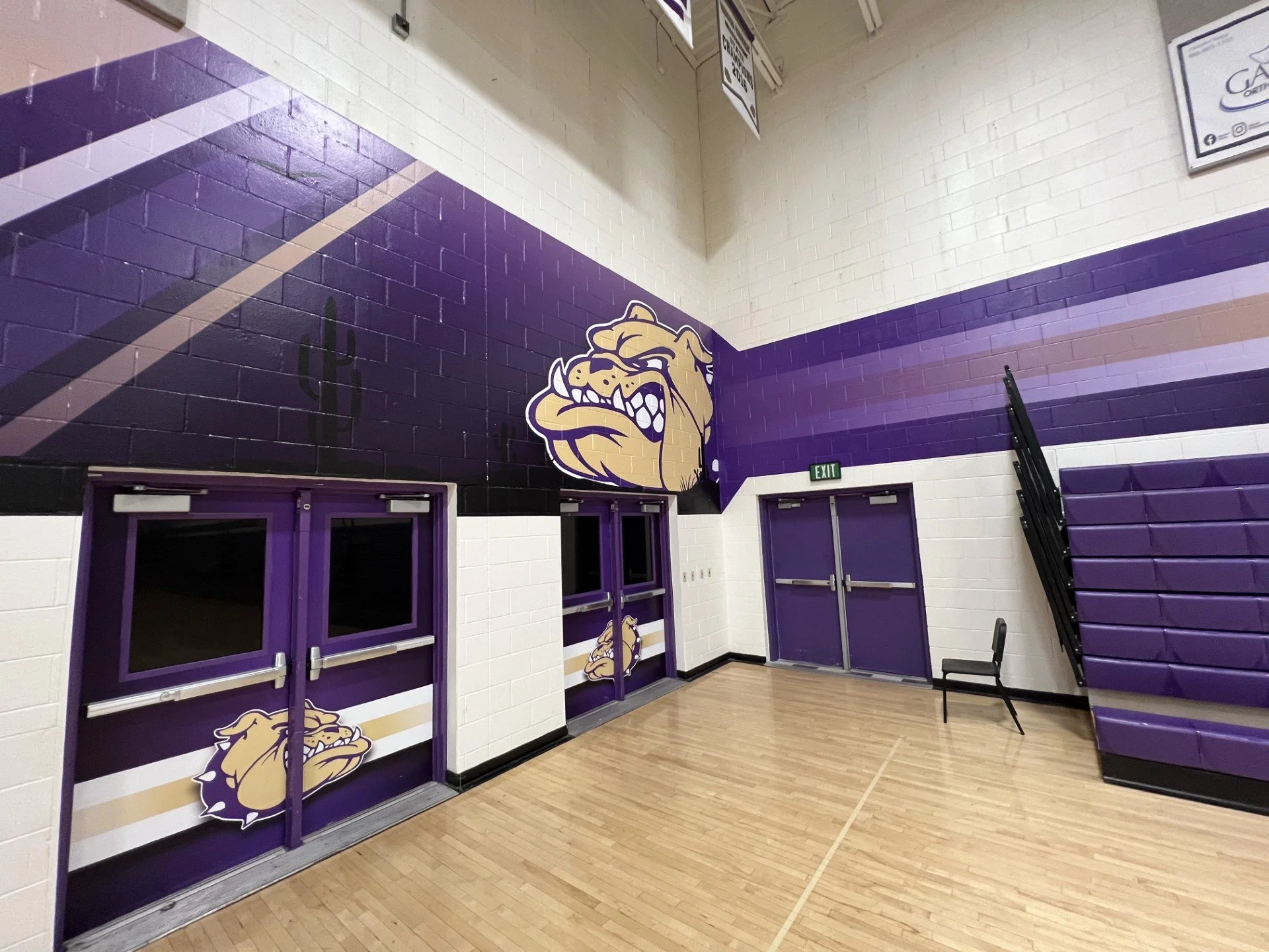 Interior of a gymnasium with purple and white walls, featuring a bulldog mascot logo, purple double doors, and purple bleachers.