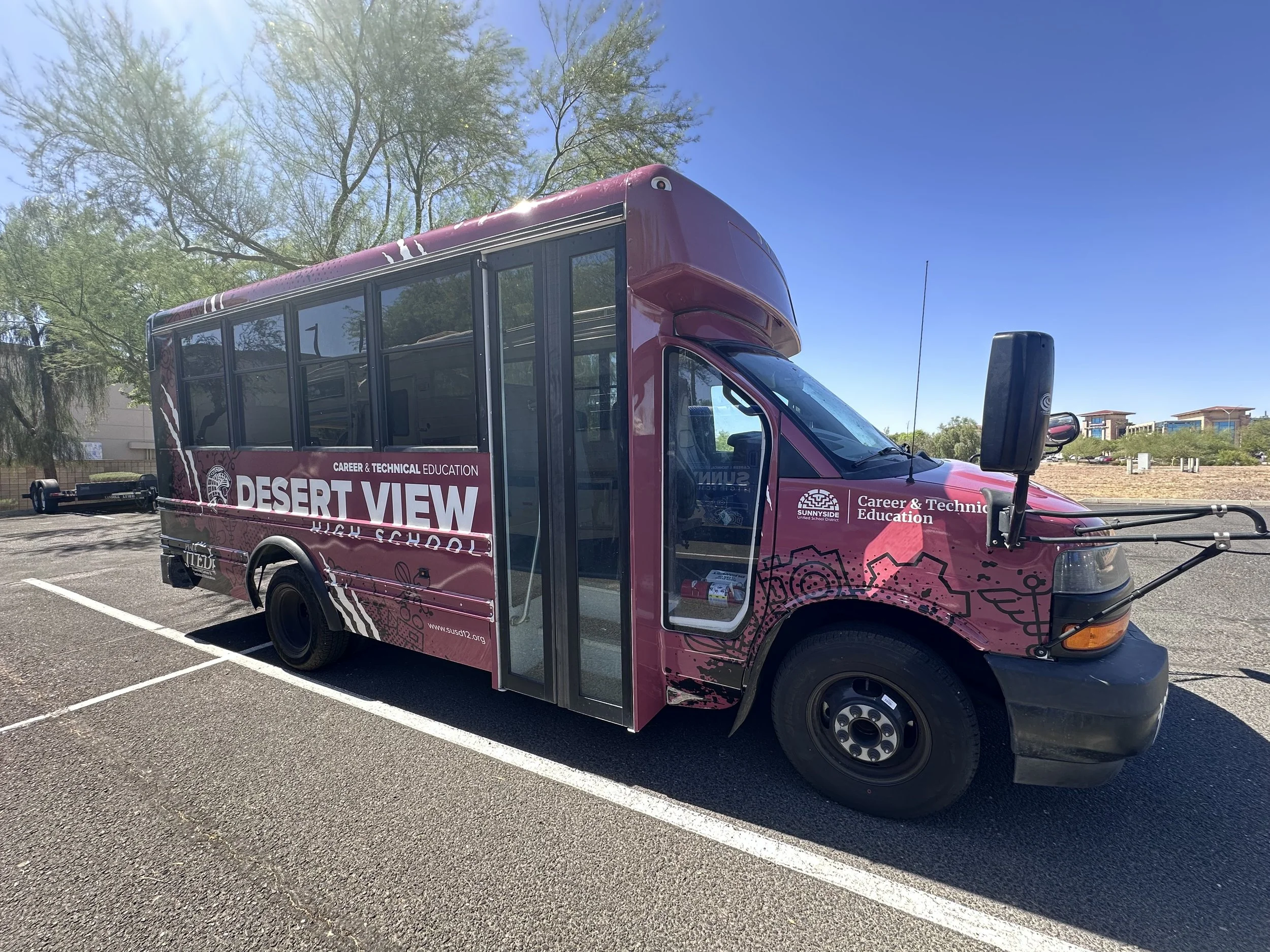 A pink shuttle bus with advertisements for career and technical education, parked in a lot with trees and buildings in the background under a clear blue sky.