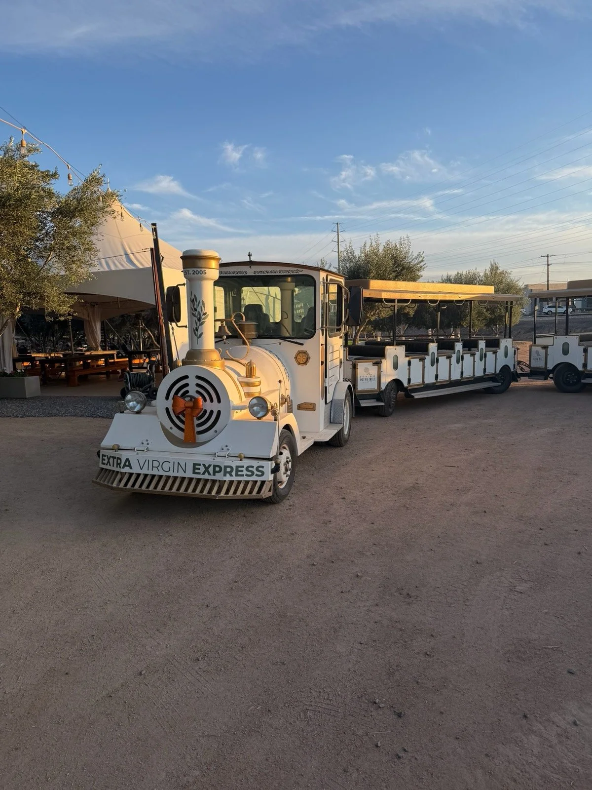 A small white tourist train with a locomotive and two train carriages, labeled "Extra Virgin Express," parked on a dirt area during sunset. There is a tent with picnic tables and trees in the background.