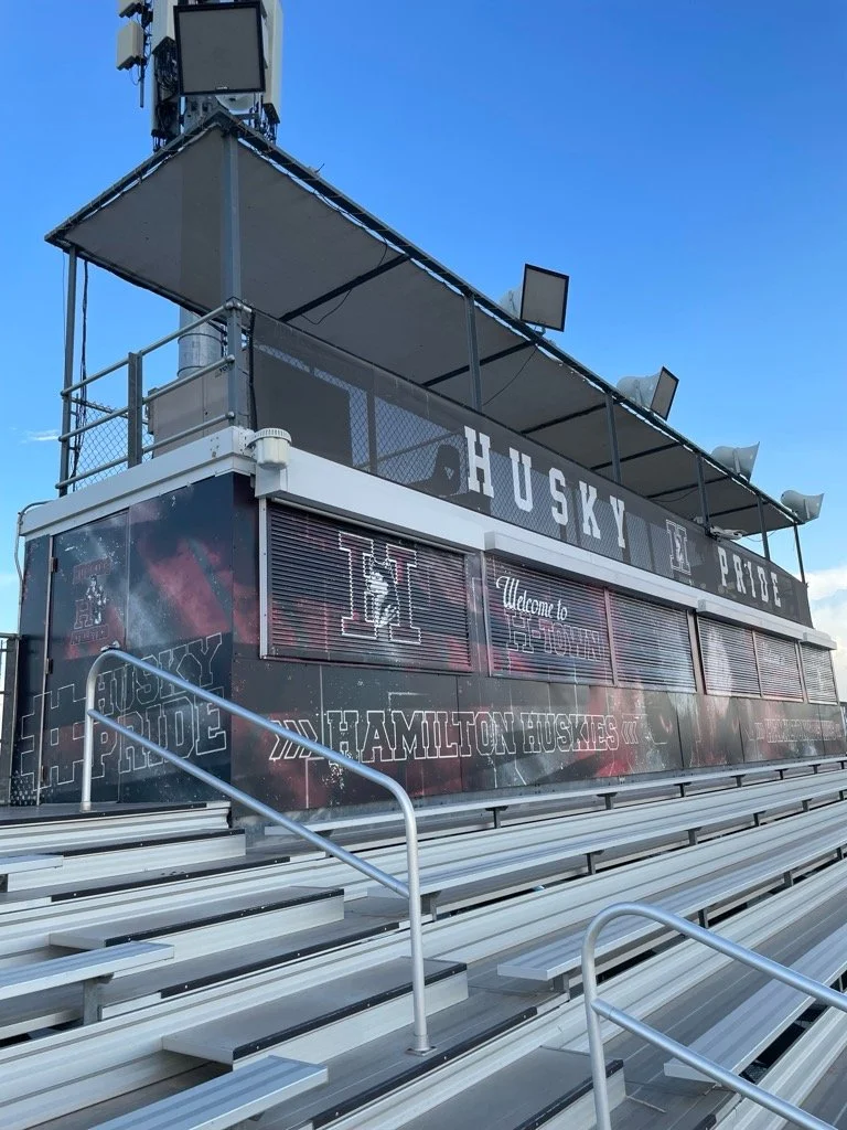 Empty outdoor football stadium bleachers with a press box and scoreboard displaying 'HUSKY PRIDE' and 'WELCOME TO HAMILTON HUSKIES' under a clear blue sky.