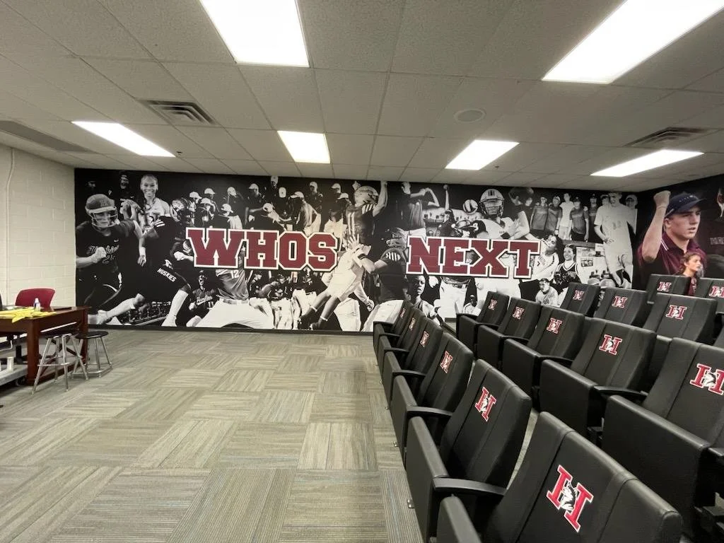 A room with black theater seats labeled with an 'H' logo and a large wall mural of black and white sports action photos, with 'WHO'S NEXT' written in bold red and white letters.