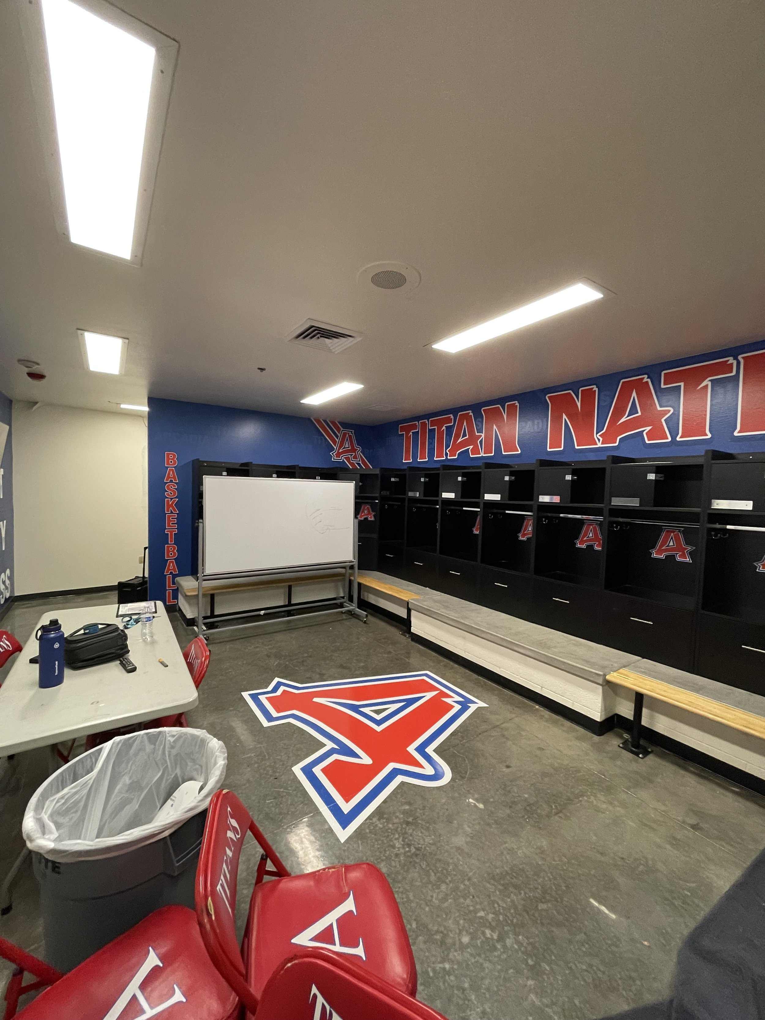 Locker room with black lockers decorated with the University of Arizona Wildcats Logo, a whiteboard, benches, red chairs with white Alpha Tau Omega lettering, and a large Arizona Wildcats logo on the floor.