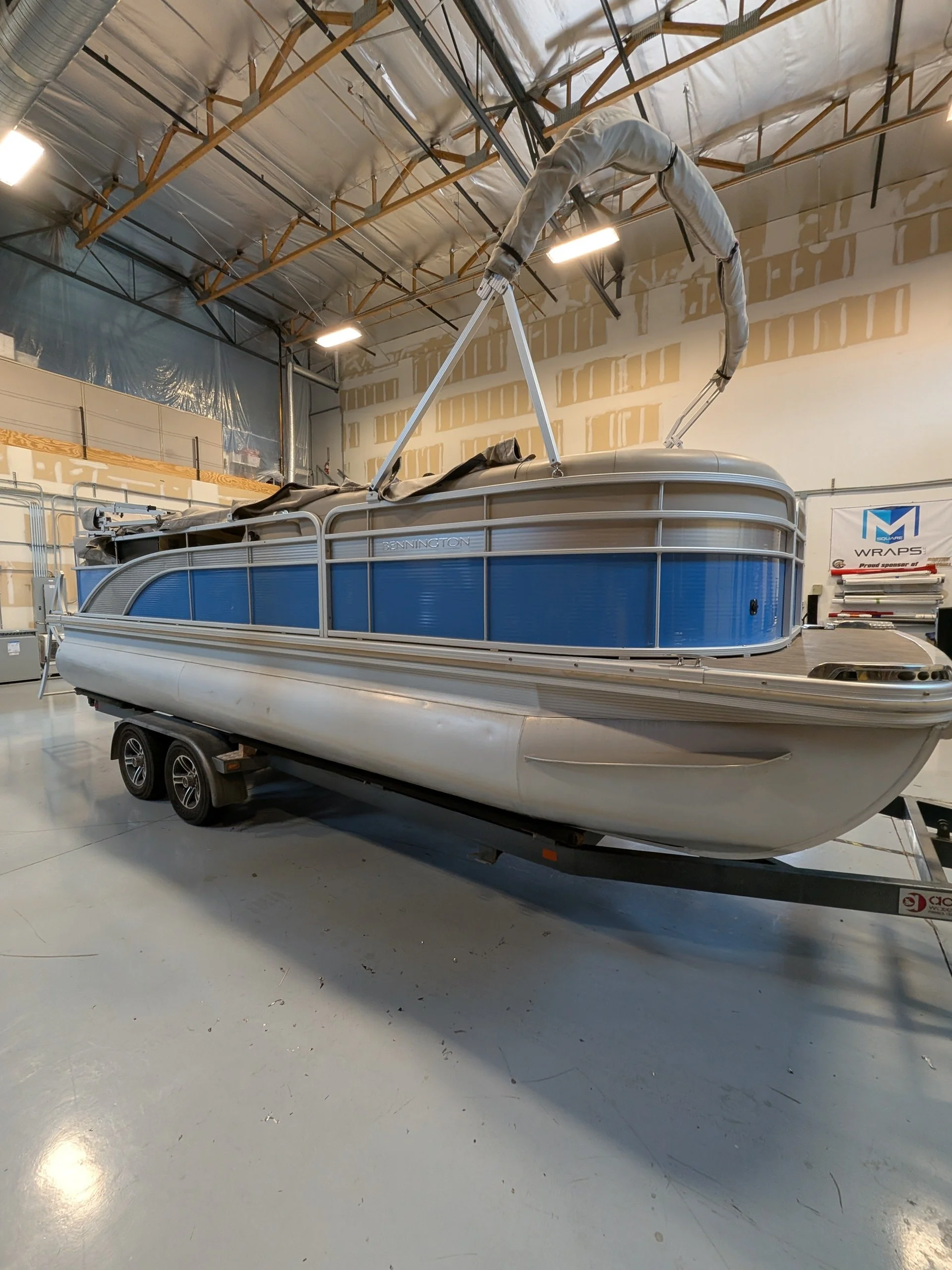 An enclosed pontoon boat on a trailer inside a warehouse with unfinished walls and ceiling.