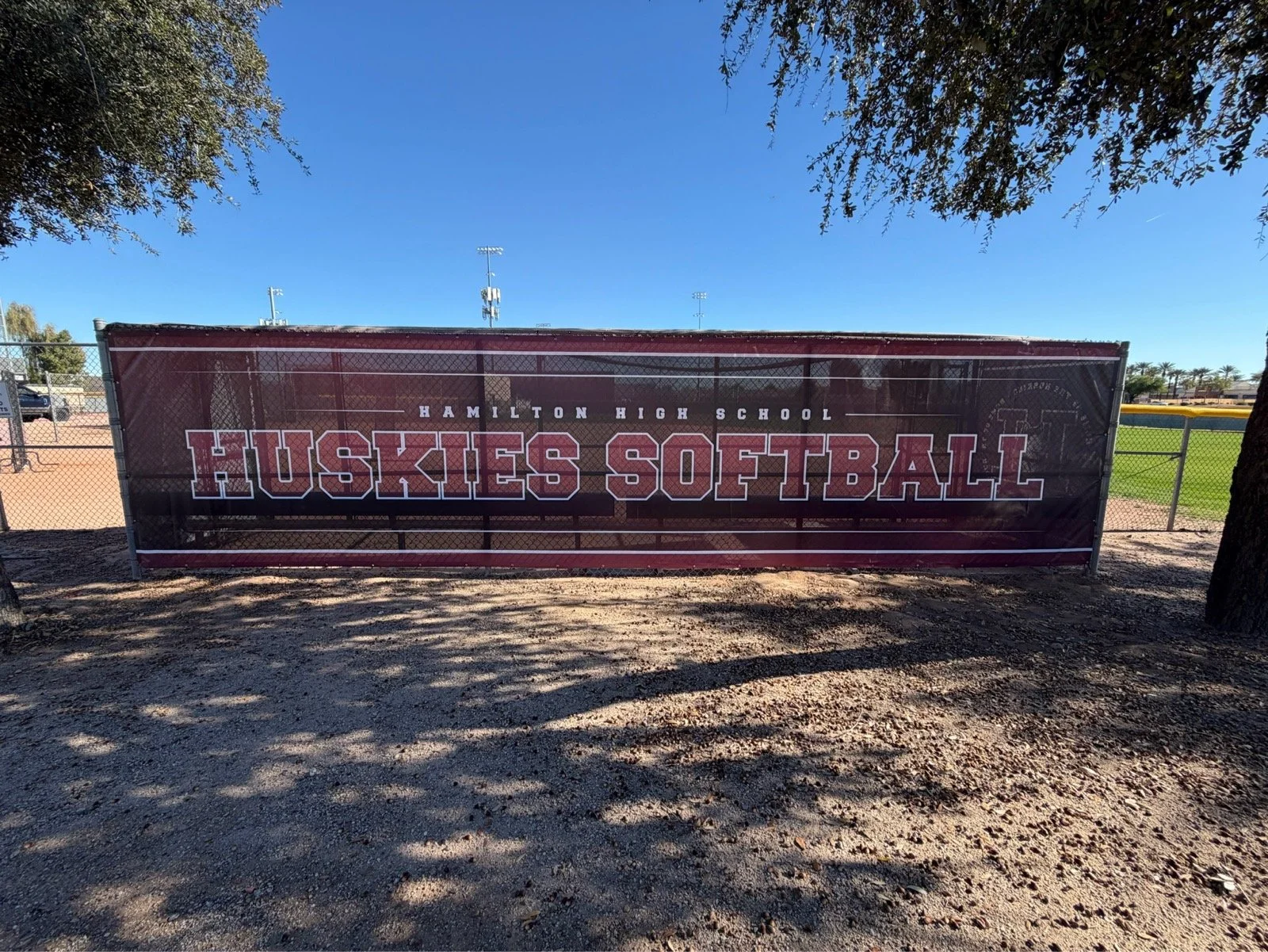 Printed sign reading "Hamilton High School Huskies Softball" on a fence at a high school softball field, with a clear blue sky and trees in the background.