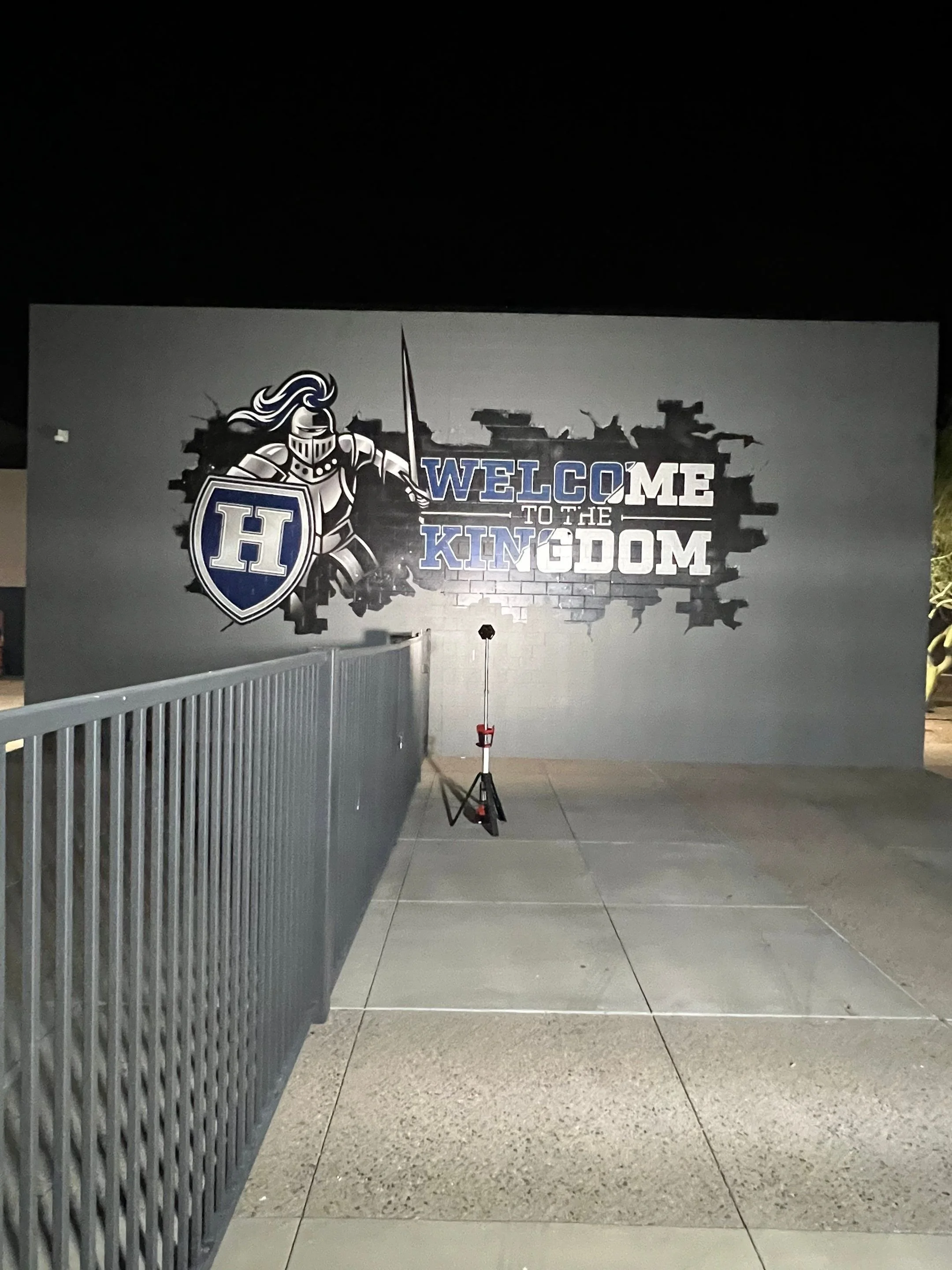 Nighttime scene of a large wall painting featuring a knight in armor holding a shield with a letter H. The text 'Welcome to the Kingdom' is painted on the wall, with a brick wall illustration behind it. There is a metal railing on the left side and a