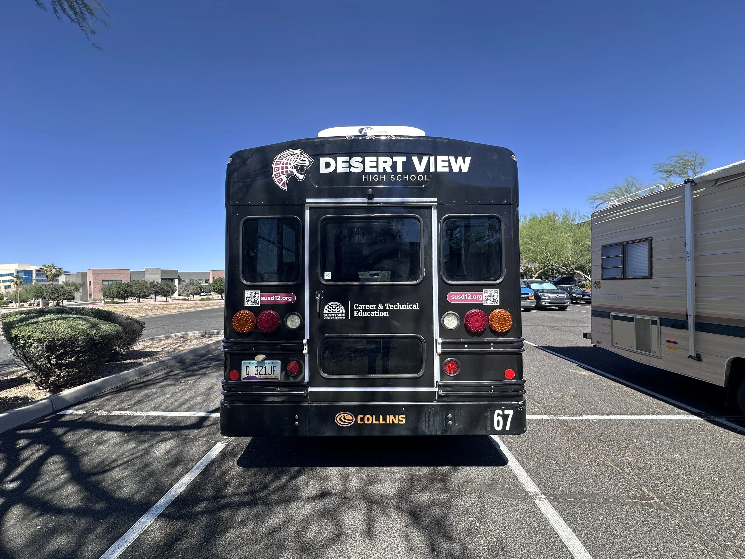 The back of a black school bus labeled 'Desert View High School' parked in a parking lot with some trees, other vehicles, and a building in the background under a clear blue sky.