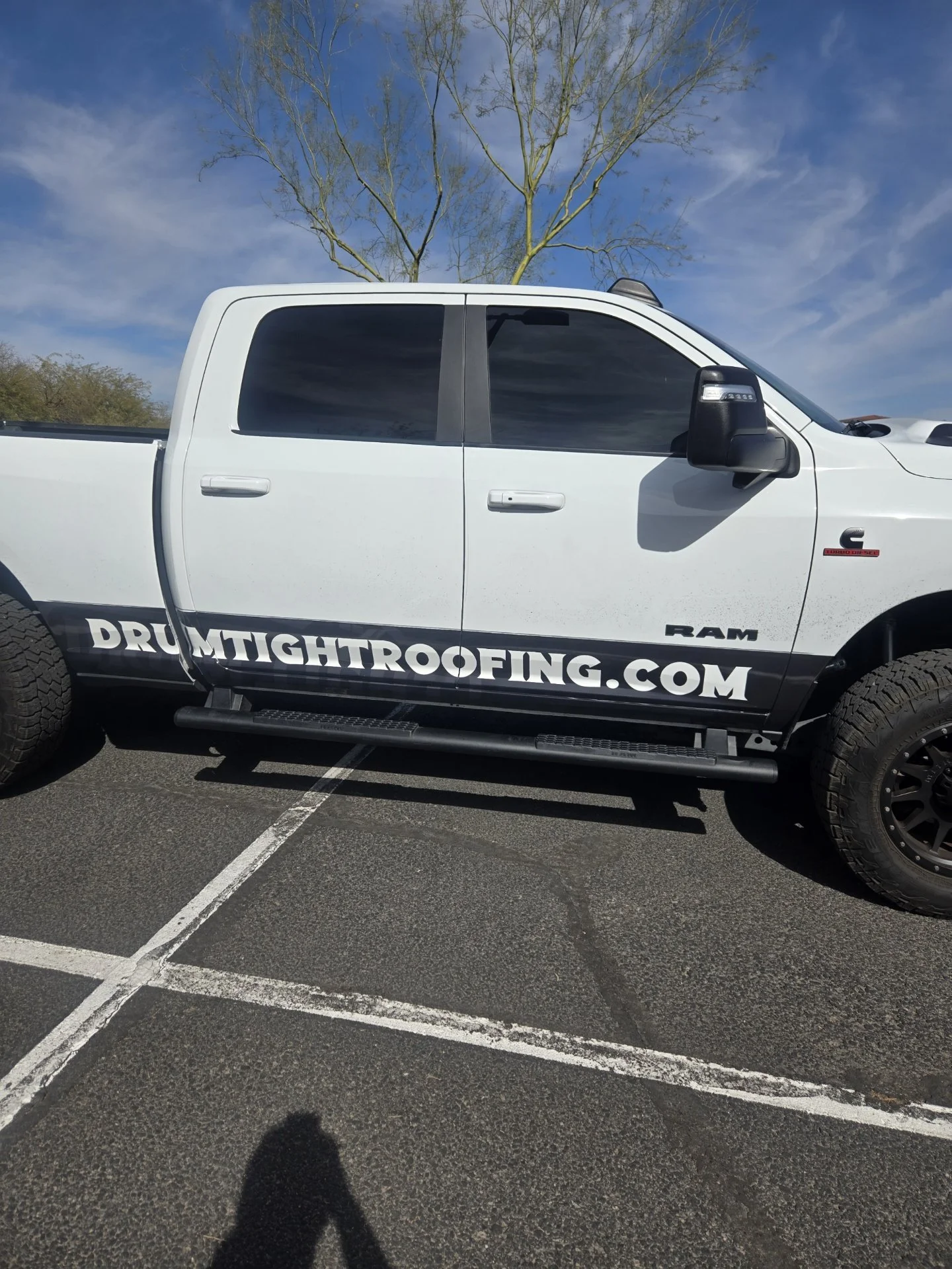 White pickup truck with black wheels and tinted windows parked in a lot with painted parking lines, under a blue sky with scattered clouds and a tree in the background. The truck has a decal on the side that reads "DRUMTIGHTROOFING.COM".