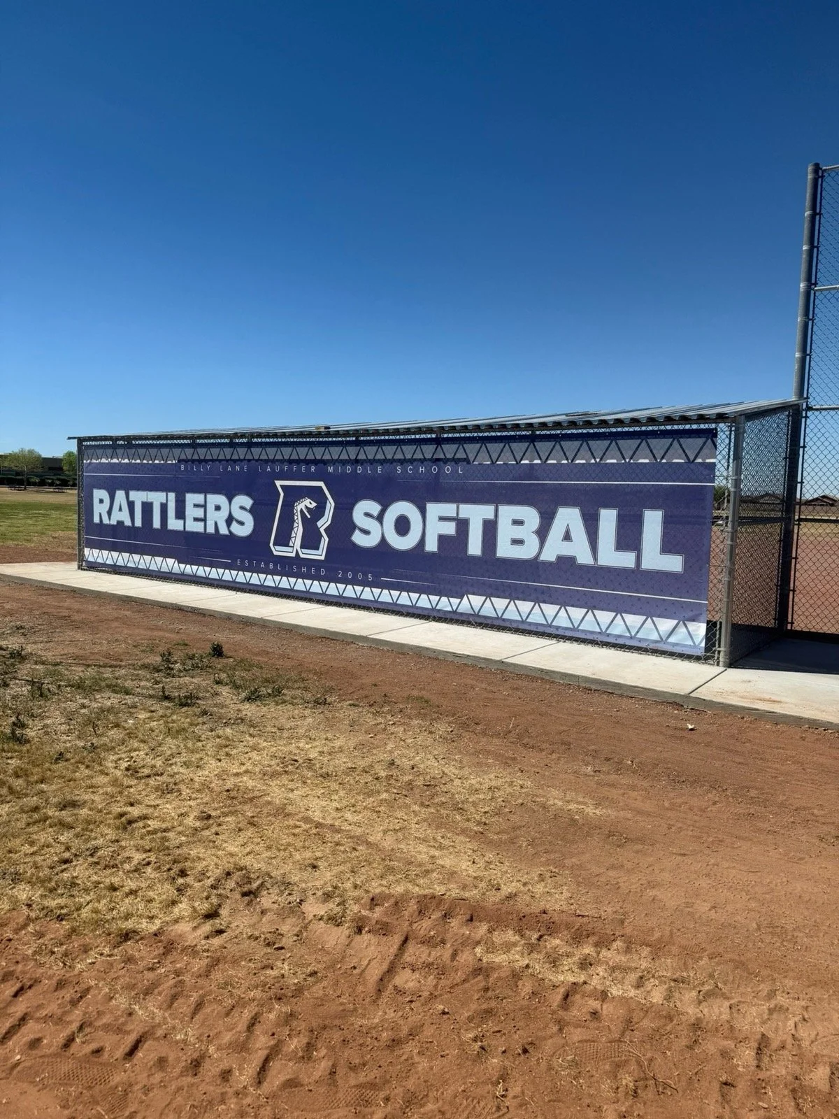 Sign for Billy Lane Lauffer Middle School Rattlers softball team, established in 2005, mounted on a chain-link fence next to a baseball field.