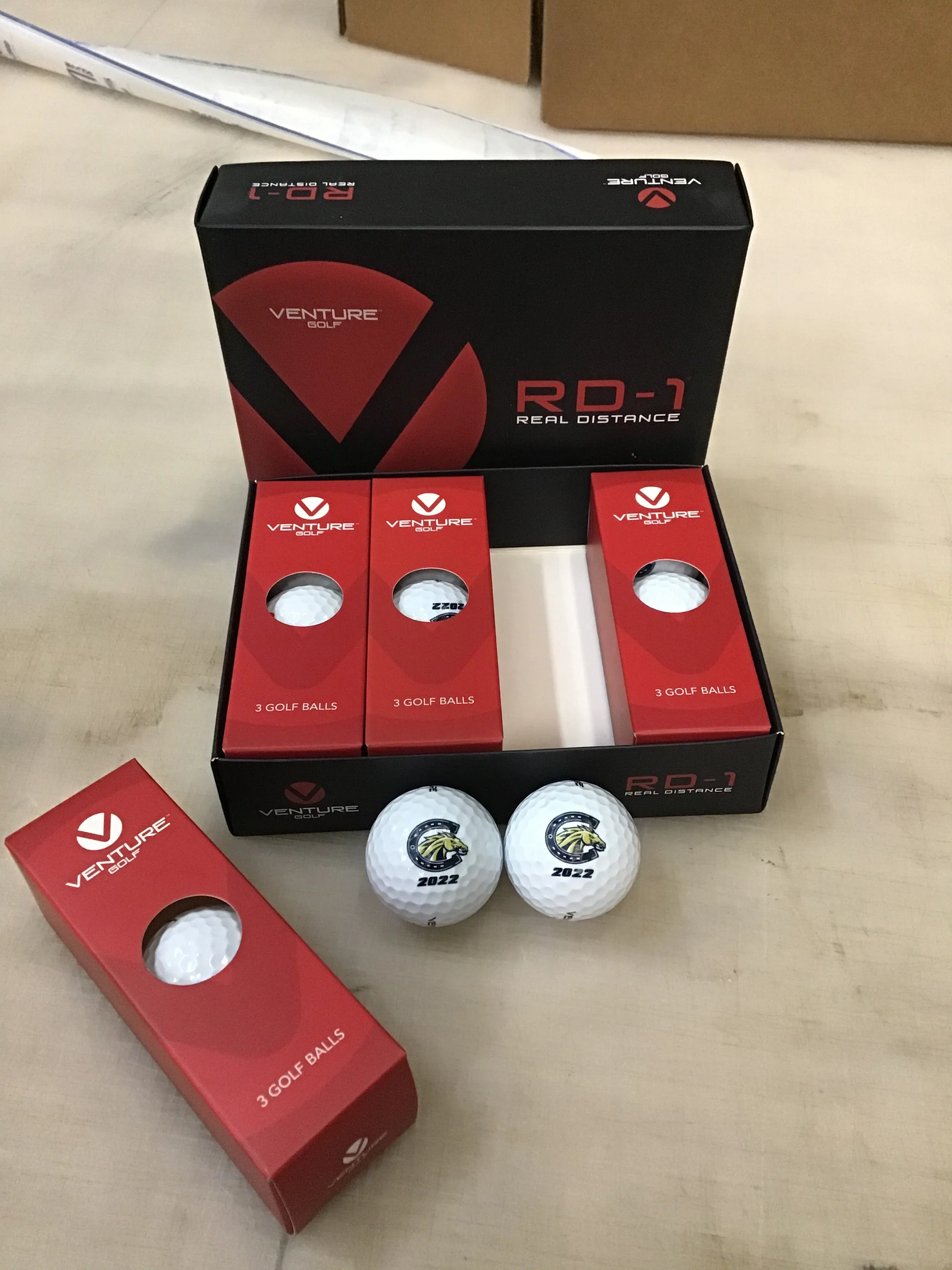 Boxes of Venture Golf balls and two golf balls with a lion logo and the year 2022 printed on them, arranged on a table.