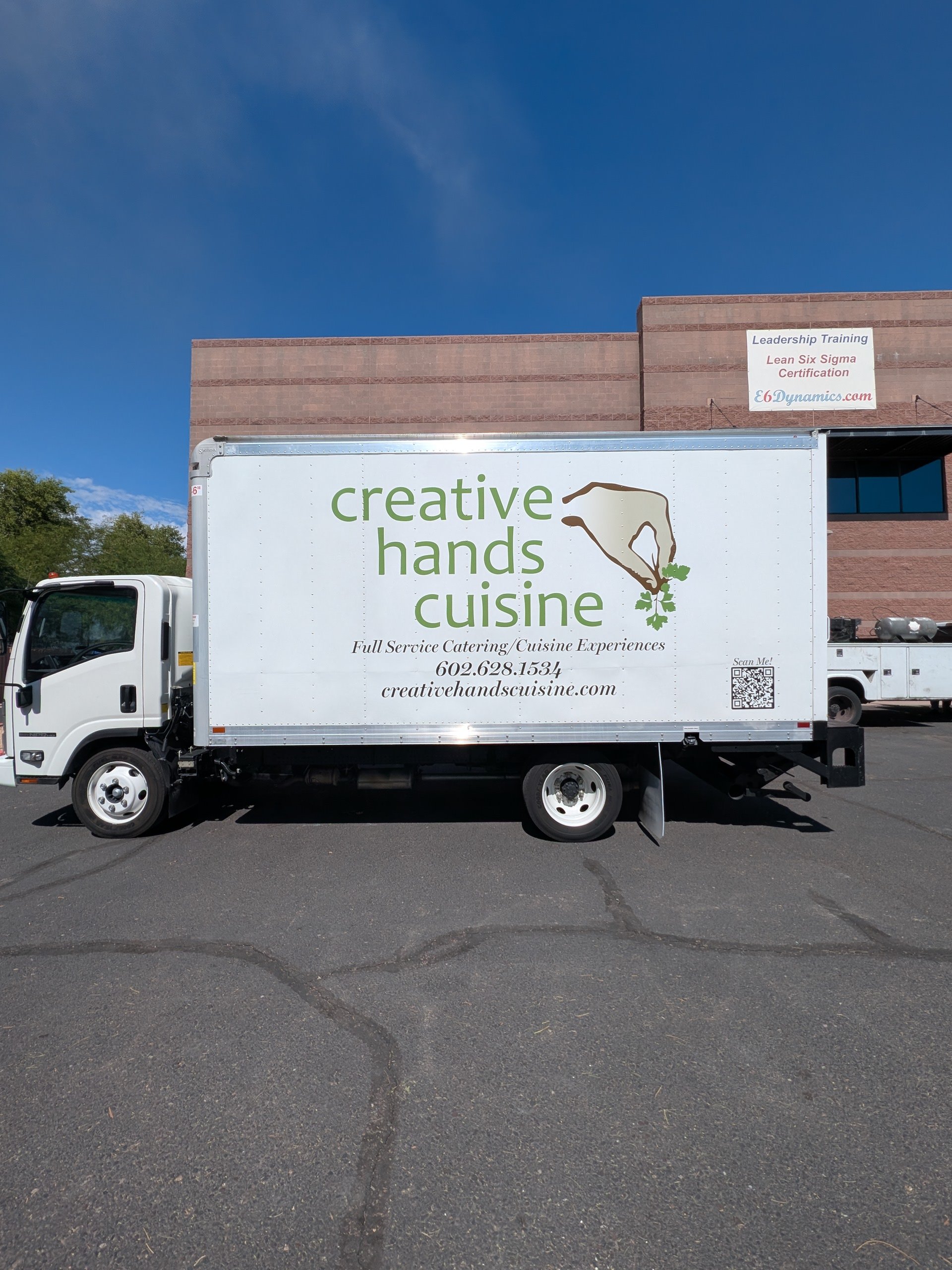A white delivery truck with a sign that reads 'creative hands cuisine' parked on a paved lot, with a brick building and blue sky in background.