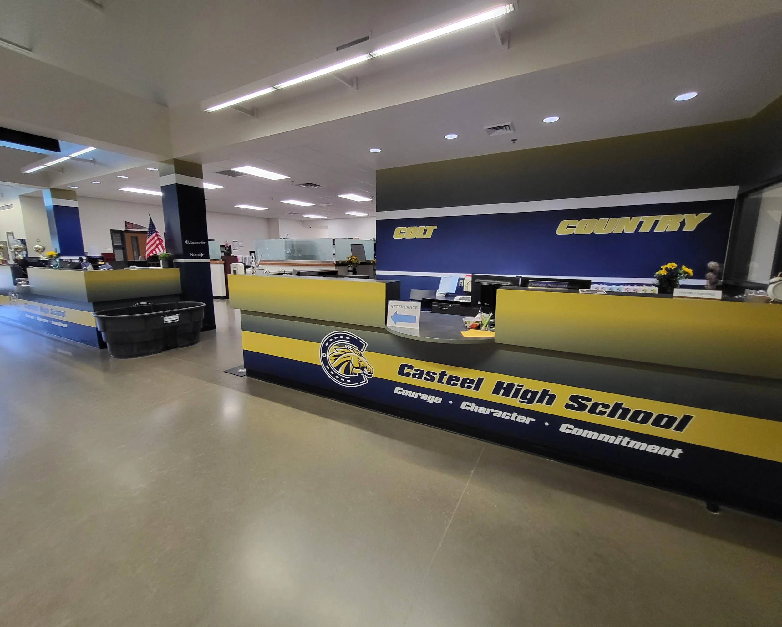 Interior view of a school office reception area with a blue and yellow school banner reading 'Castlea High School' and a counter with a computer and flowers, with a sign pointing towards attendance.