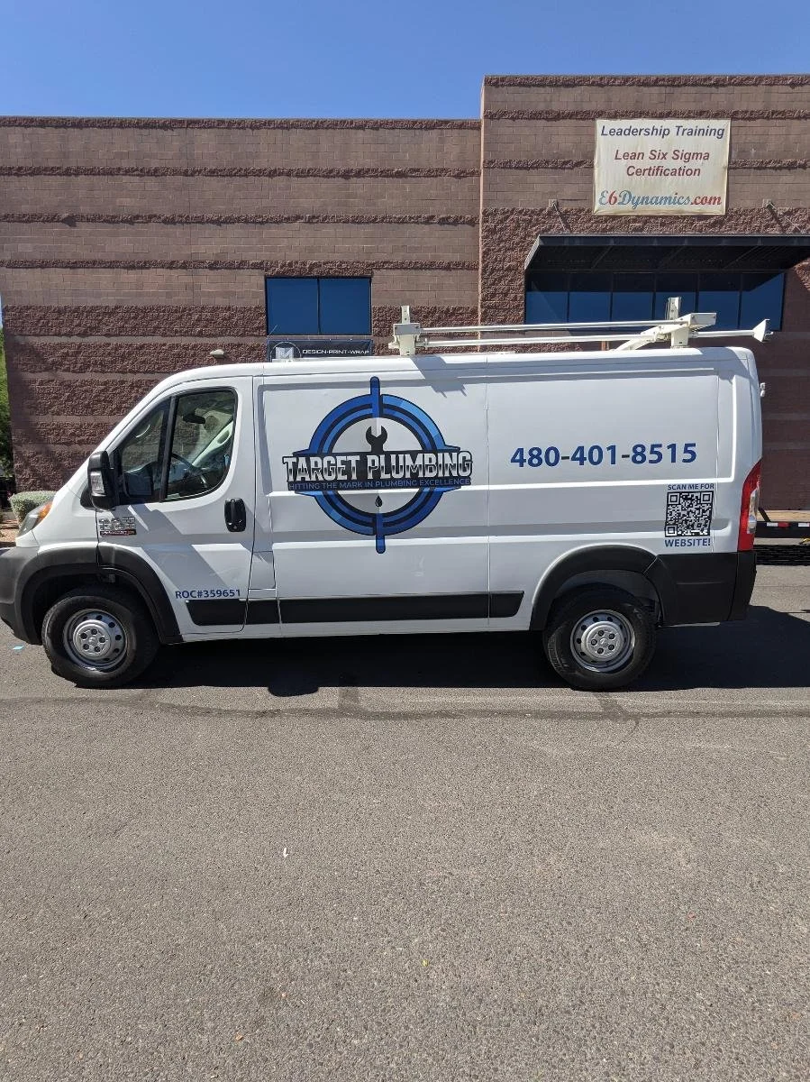 A white service van for Target Plumbing parked in front of a brick building with a sign that reads 'Leadership Training Lean Six Sigma Certification.' The van displays the company's logo, phone number, a QR code for the website, and the text 'TRADING